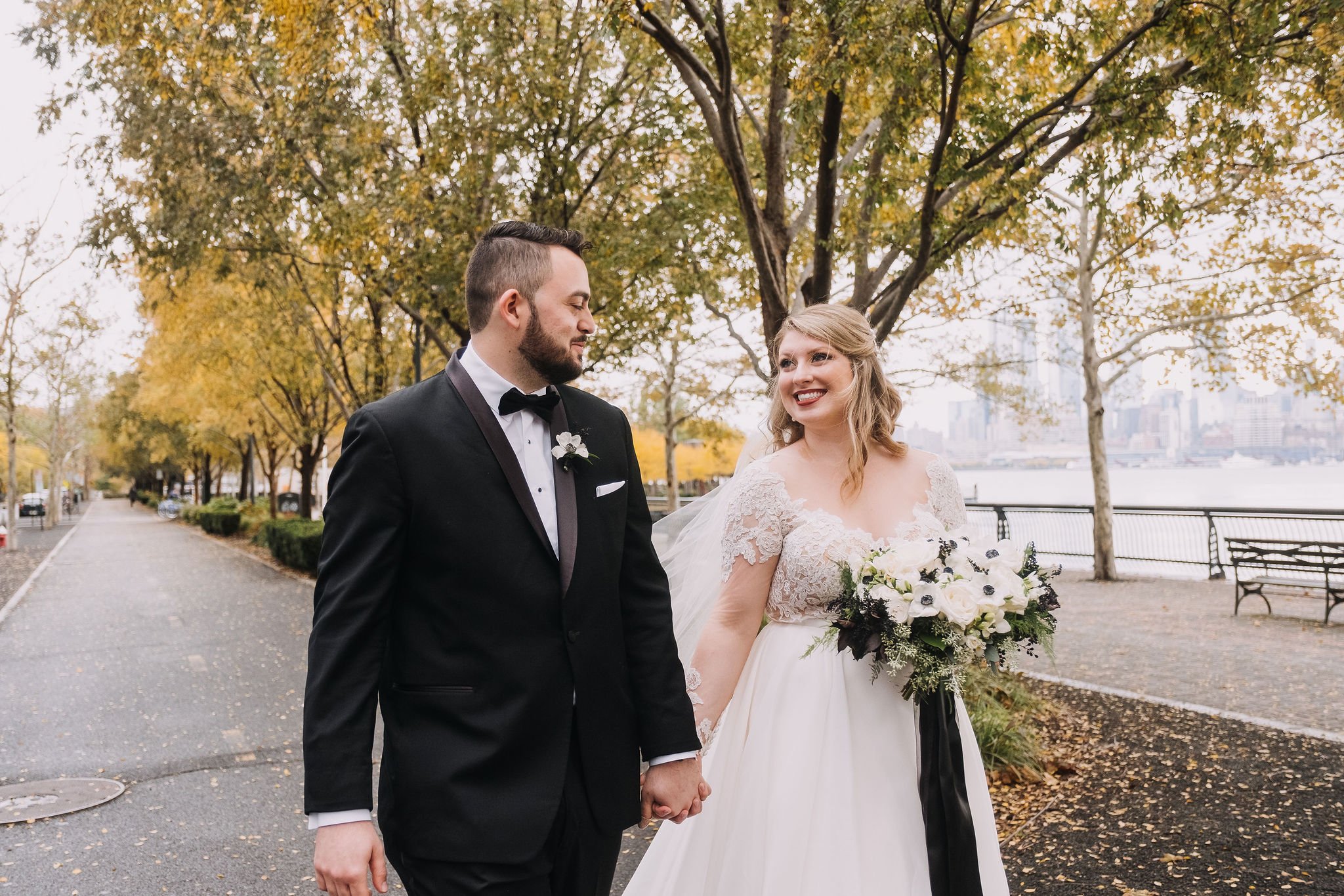 A bride and groom holding hands outdoors in a park, with autumn foliage and city skyline in the background. The groom is dressed in a black tuxedo, and the bride is wearing a white wedding gown and holding a bouquet of white and dark flowers.