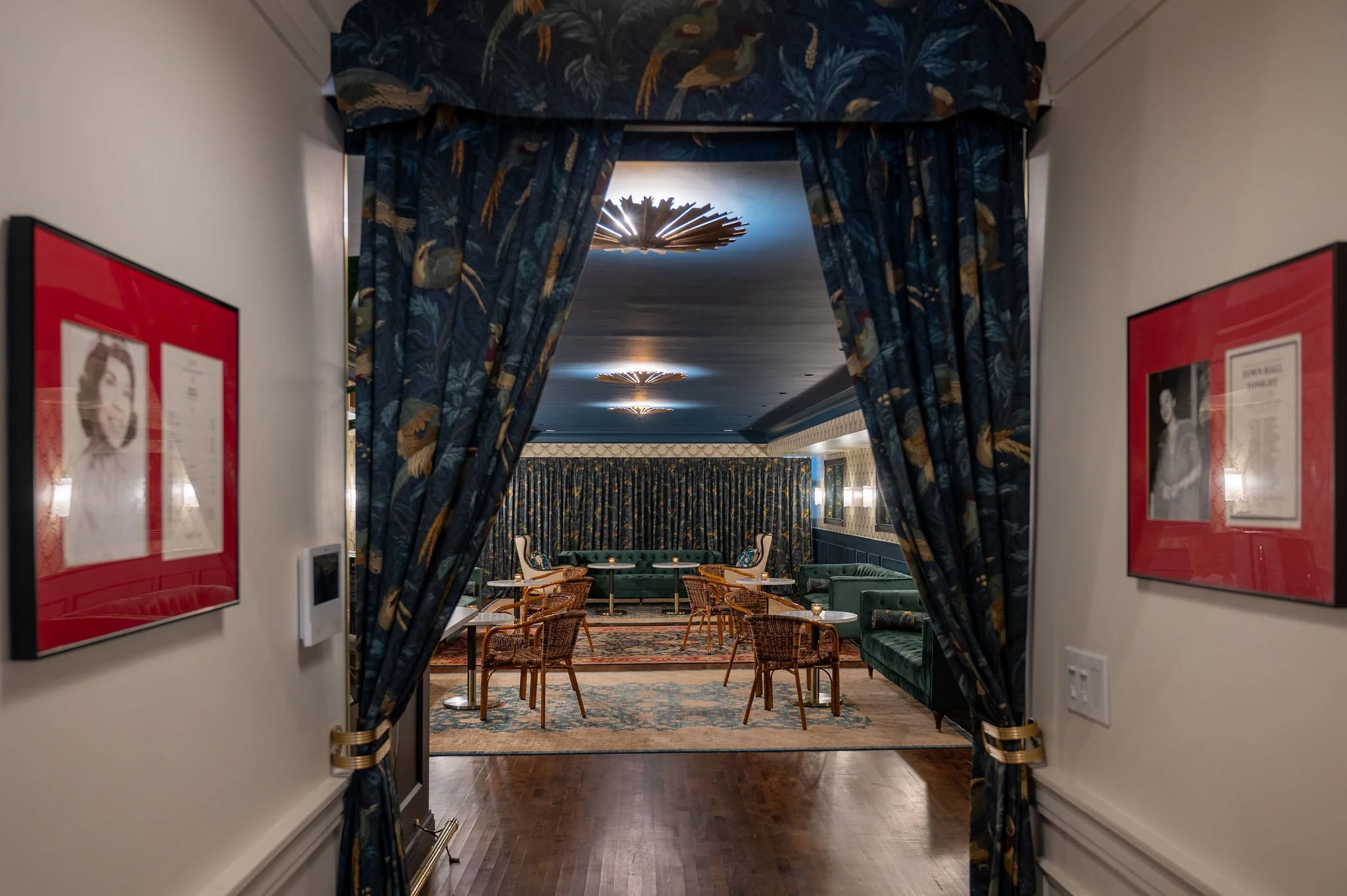 A view of a hotel or restaurant interior seen through a doorway with dark blue patterned curtains. The room features a seating area with green velvet sofas, wooden chairs, tables, and ornate rugs. The ceiling has decorative lighting fixtures.