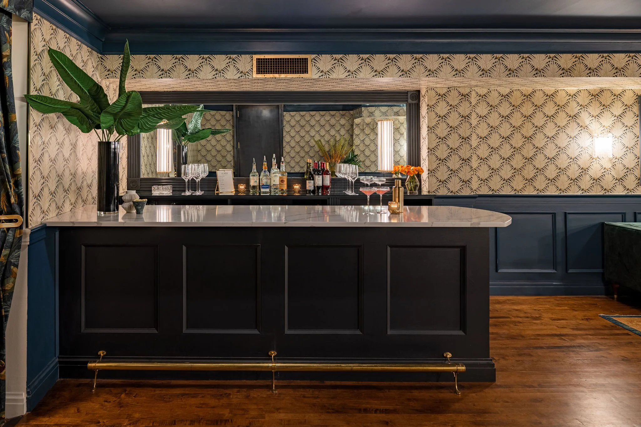 A bar counter with liquor bottles, glasses, and a menu, decorated with large houseplants and floral arrangements, set against wallpaper with a leaf pattern and framed mirrors, in a stylish interior.