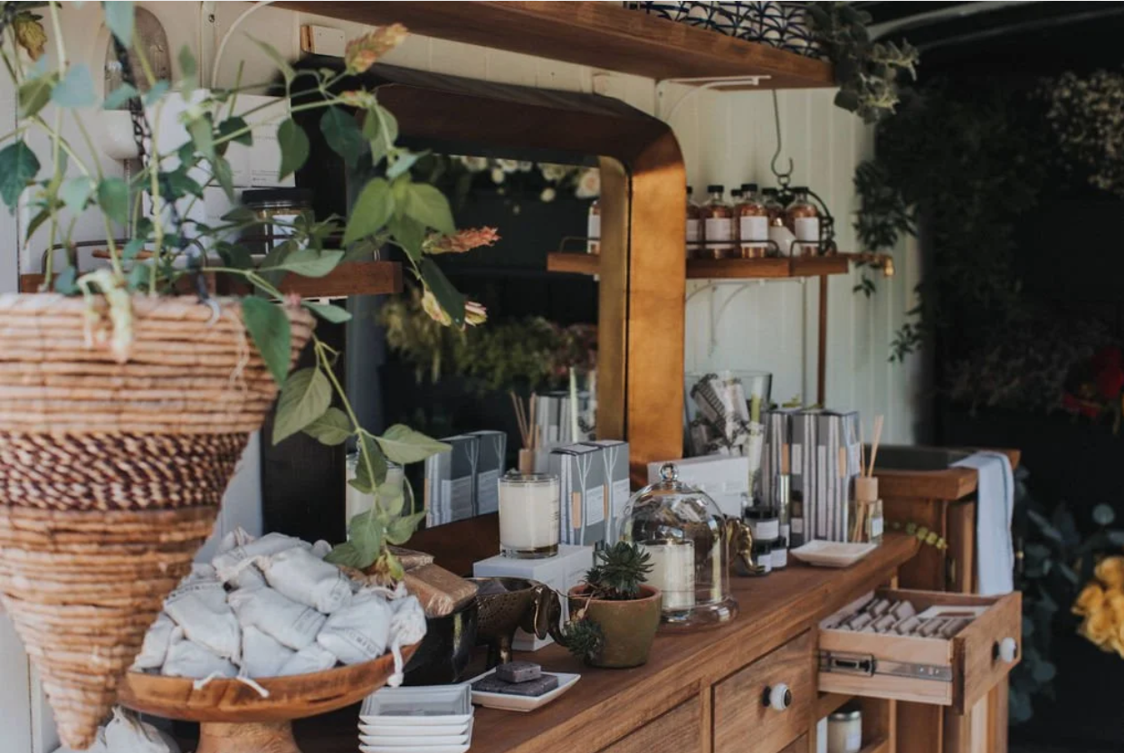 Wooden vanity table with various skincare and cosmetic products, potted plants, candles, and decorative items, with a mirror and shelves in the background.