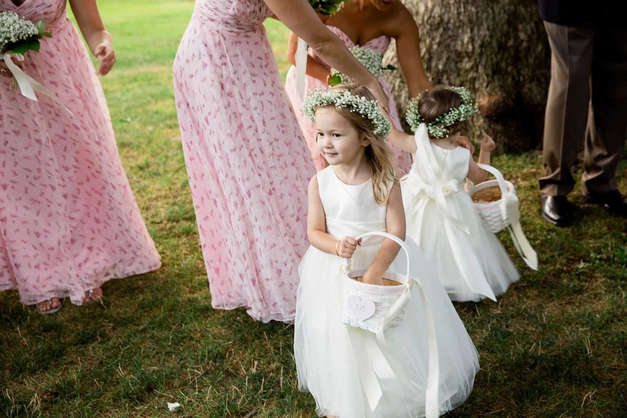 Young girls in white dresses and floral crowns participating in a wedding celebration, holding baskets, with women in pink dresses and bouquets nearby, outdoors on grass.