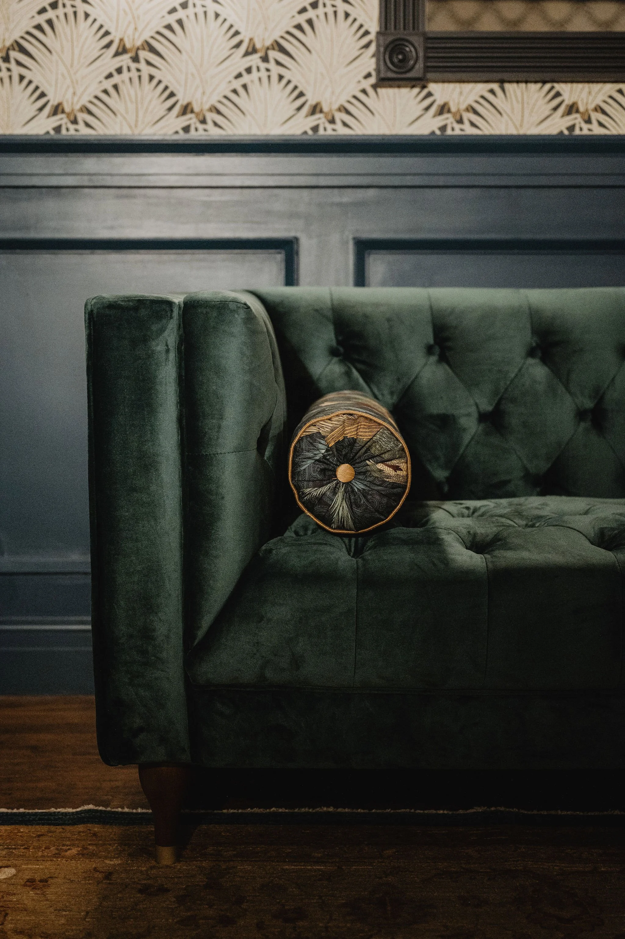 A green velvet tufted sofa with a round patterned pillow on it, in front of a dark wall with framed artwork, and a wallpapered wall with a geometric fan pattern.