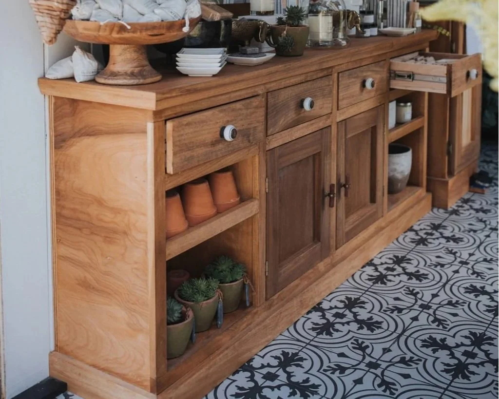 A wooden sideboard with drawers, cabinets, and open shelves, decorated with plants, pottery, and small kitchen items, on a black and white patterned tiled floor.