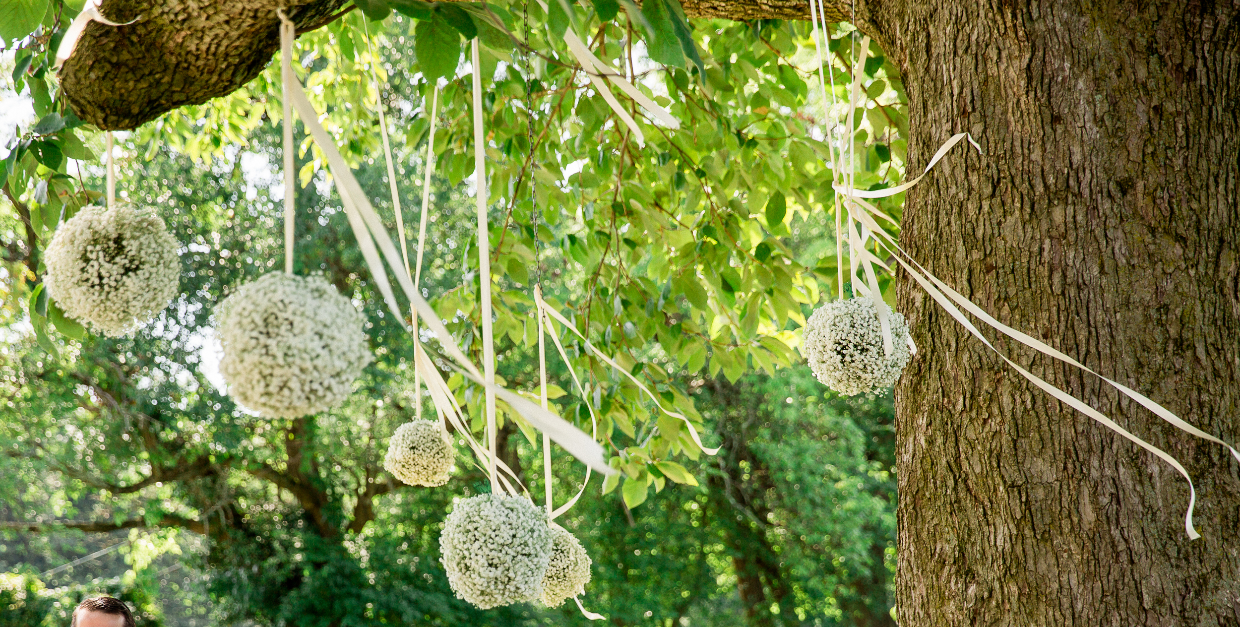 Decorative white pom-pom balls and ribbons hang from tree branches in a lush outdoor setting.