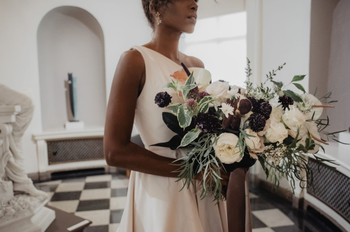Woman in a cream-colored dress holding a large bouquet of white, purple, and orange flowers with greenery in an elegant room.