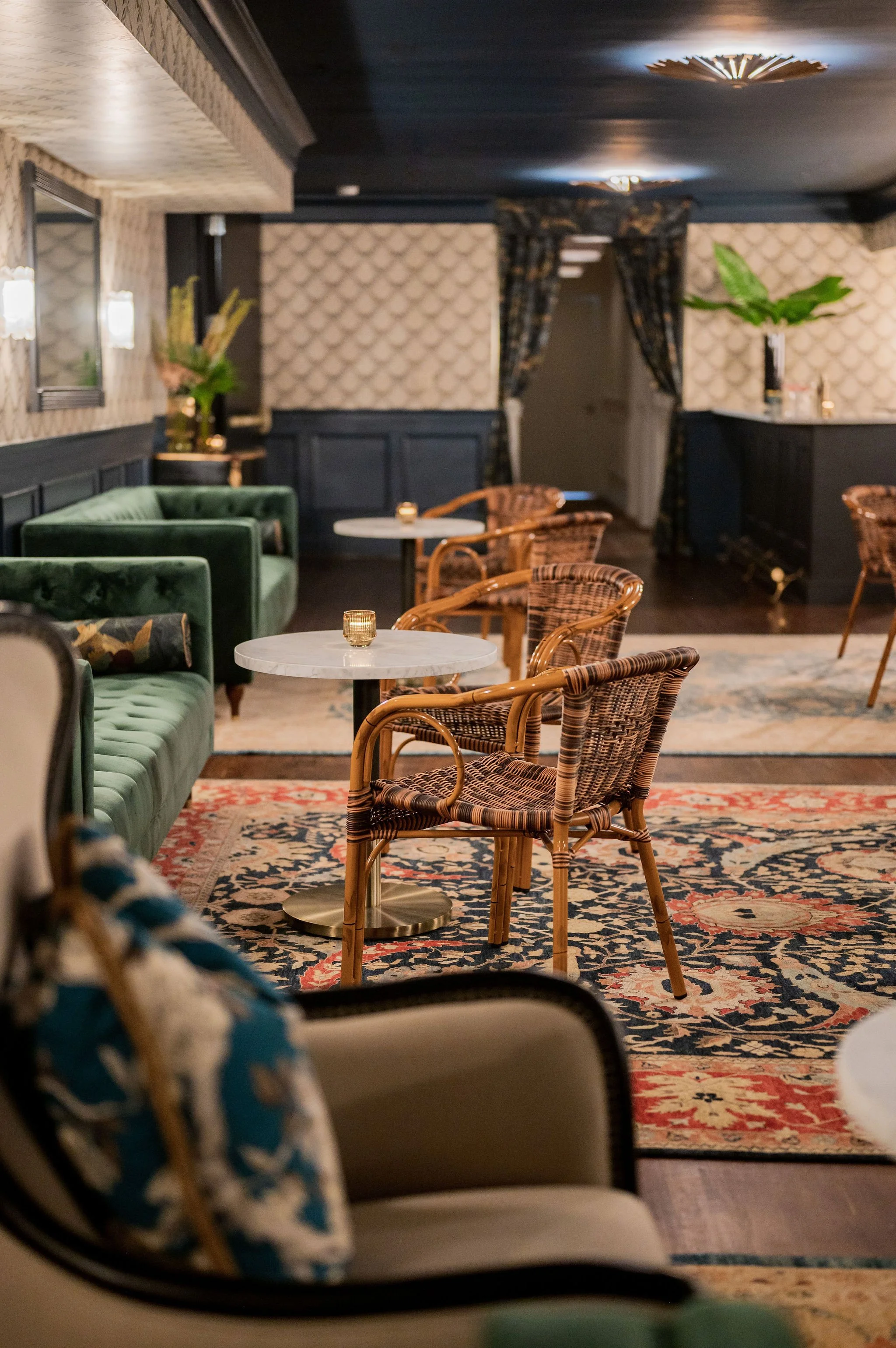 A cozy lounge area with green velvet sofas, wicker chairs, and marble-topped tables with small candles. Decor includes a patterned rug, floral arrangements, black wainscoting, and floral curtains. Dim, warm lighting creates an inviting ambiance.