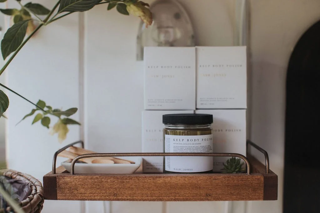 A wooden shelf with a black jar labeled 'Kelp Body Polishing,' topped with white boxes and a white plate with cookies, and some green plants nearby.