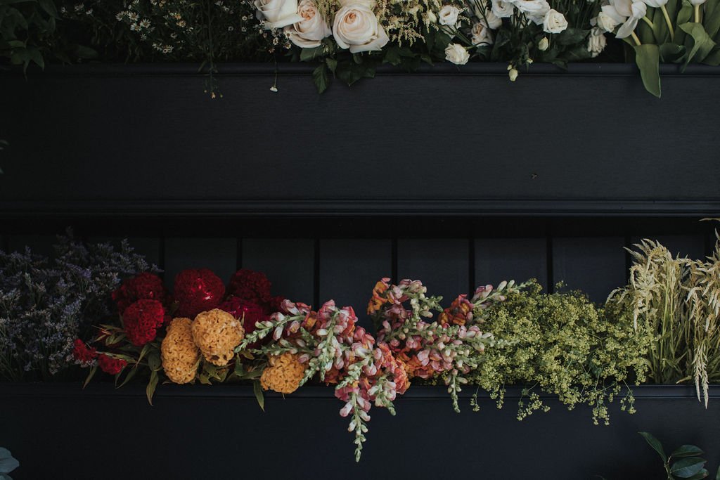 Arrangement of various flowers and greenery in black planters, including white roses, yellow and red flowers, pinkish-green sprigs, and tall ornamental grass.