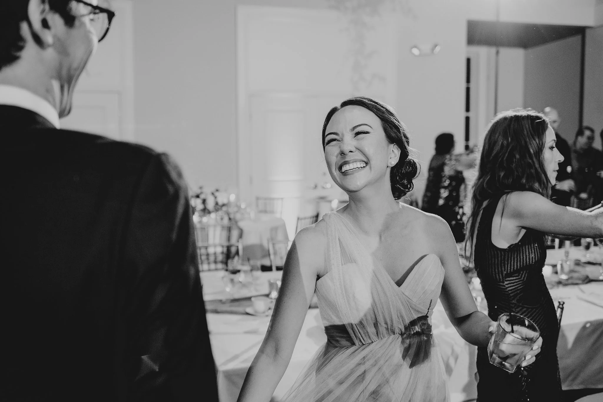 A woman in a sleeveless dress smiling and holding a drink at a social event, with other people in the background.