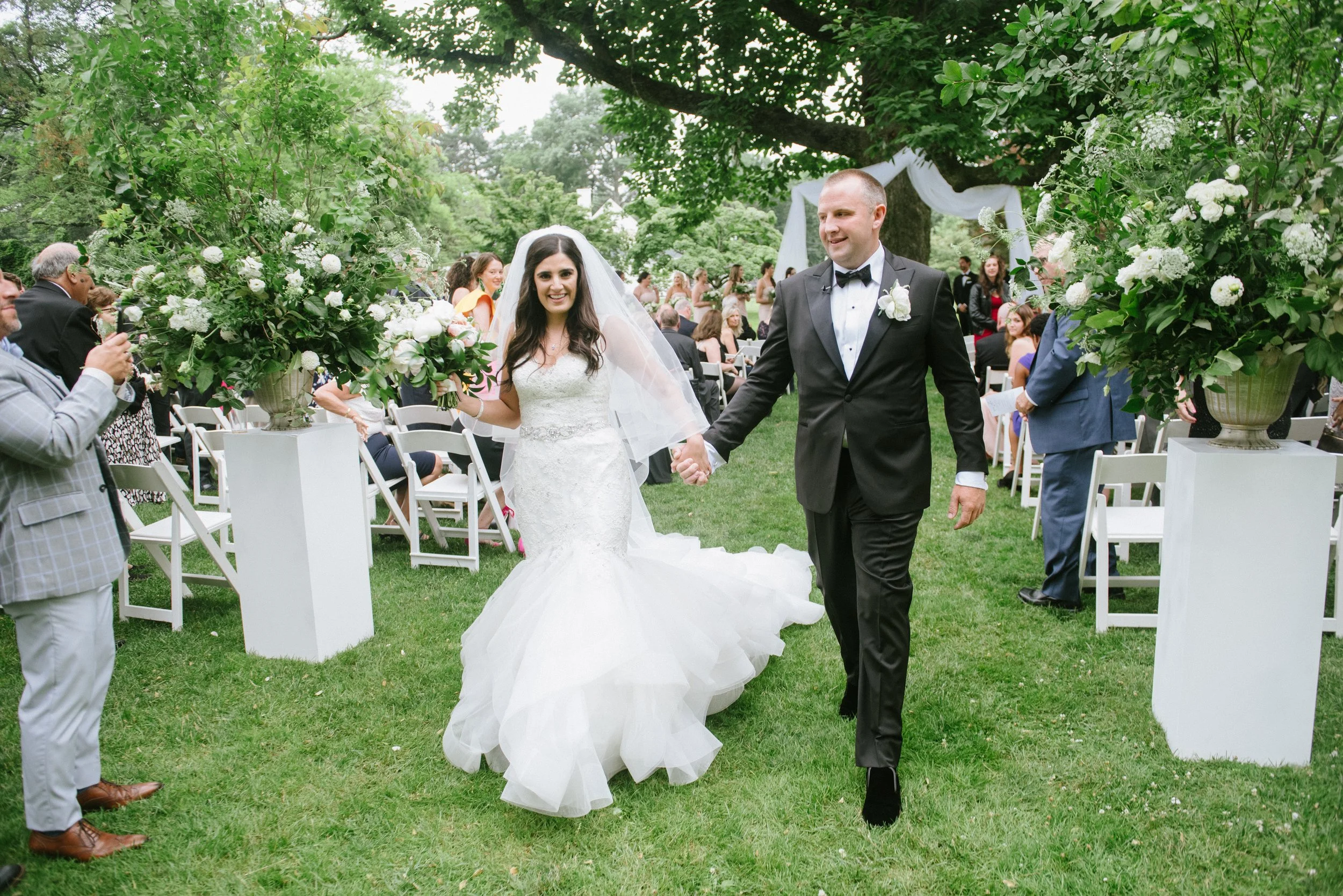 Bride and groom walking hand in hand at their outdoor wedding ceremony, surrounded by guests and floral arrangements under a large tree.
