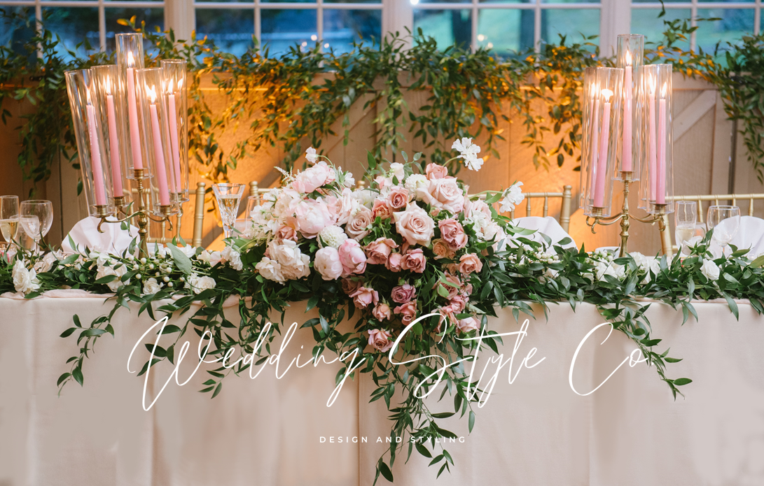Elegant wedding reception table decorated with pink roses, white flowers, lush greenery, tall pink candles in candelabras, and wine glasses at each place setting.