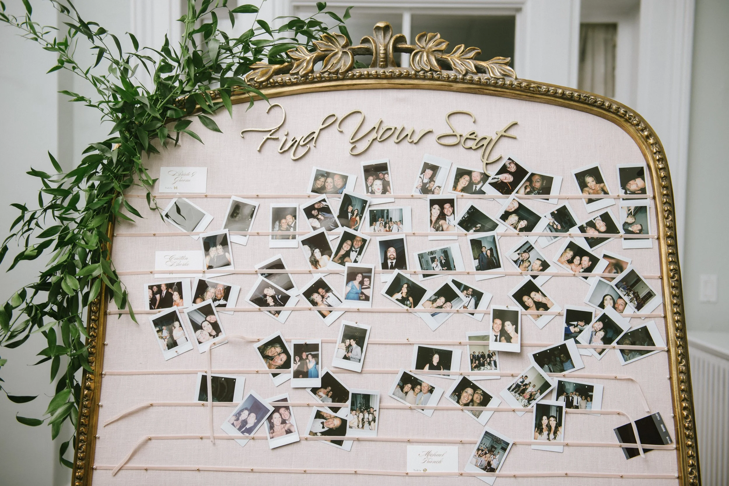 A wedding seating chart display with a decorative gold frame, green foliage, and numerous small photo prints of guests. The top reads 'Find Your Seat' in elegant script.