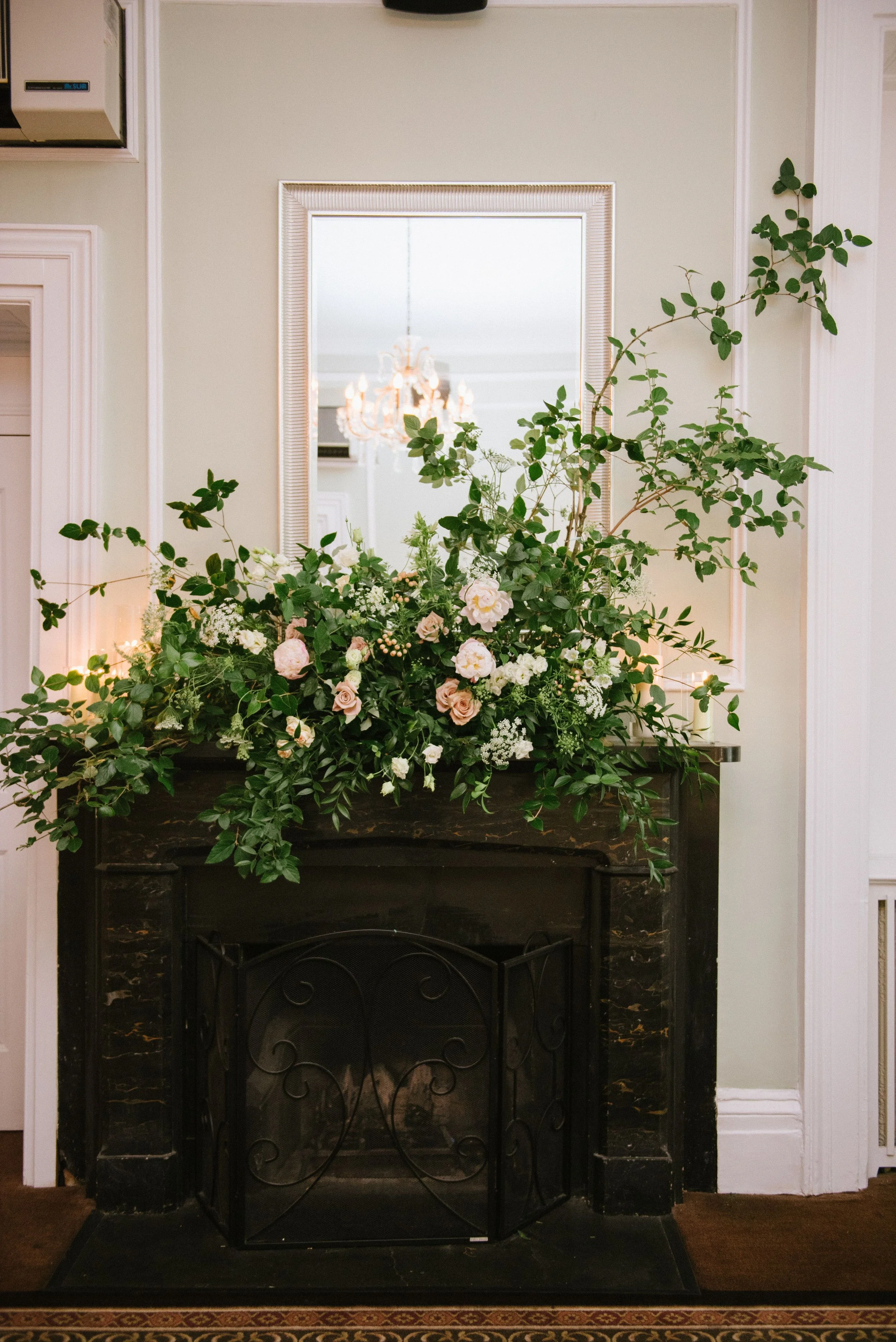 A fireplace with a dark marble surround and black metal grate, decorated with lush green foliage and pale pink and white flowers, with a mirror and chandelier reflected in the background.