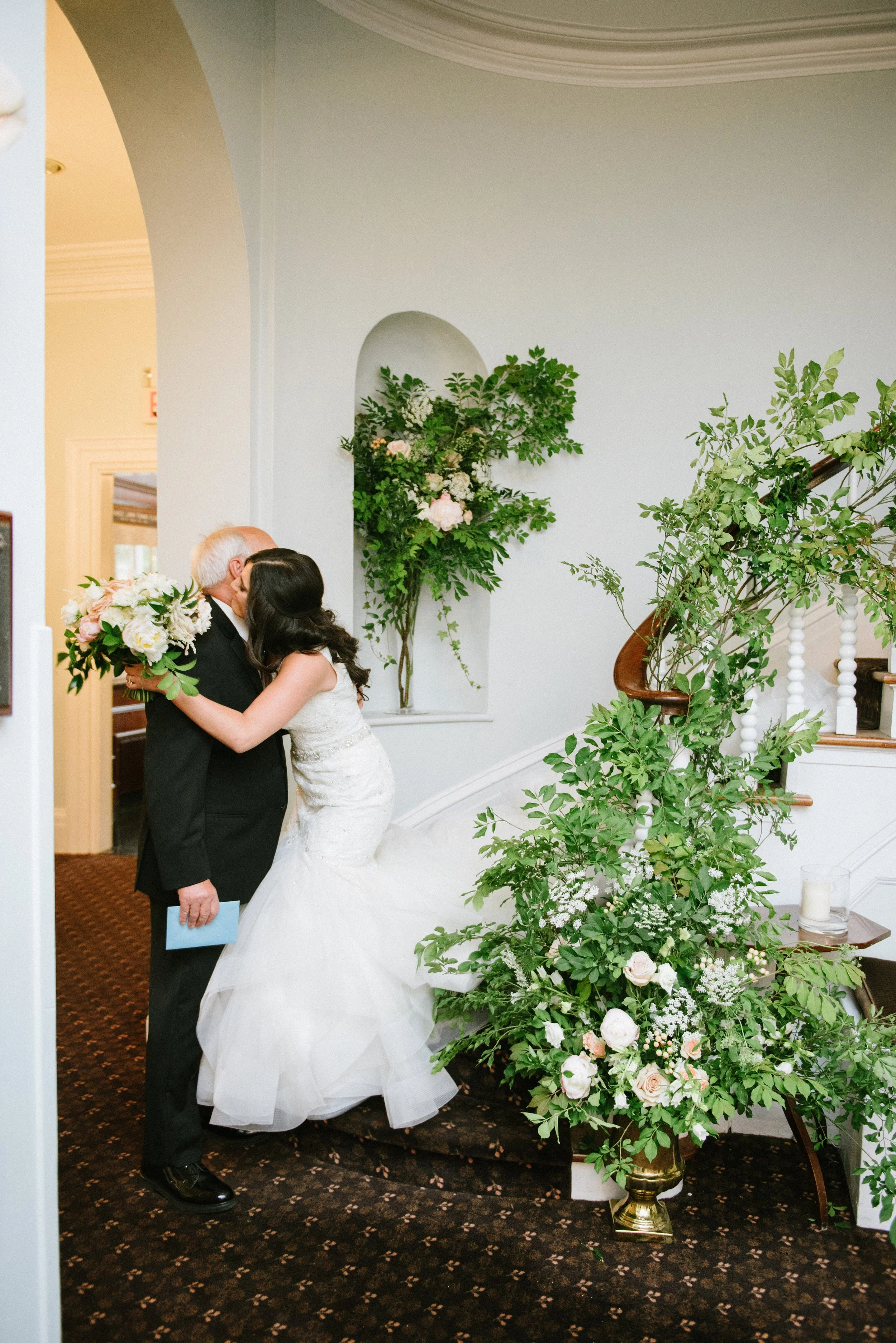 A bride and an elderly man, likely her father, kissing at a wedding ceremony. The bride is holding a bouquet of flowers, and they are standing near a floral arrangement on a staircase with greenery and pink and white flowers. The setting is inside an elegant building with white walls and decorative molding.
