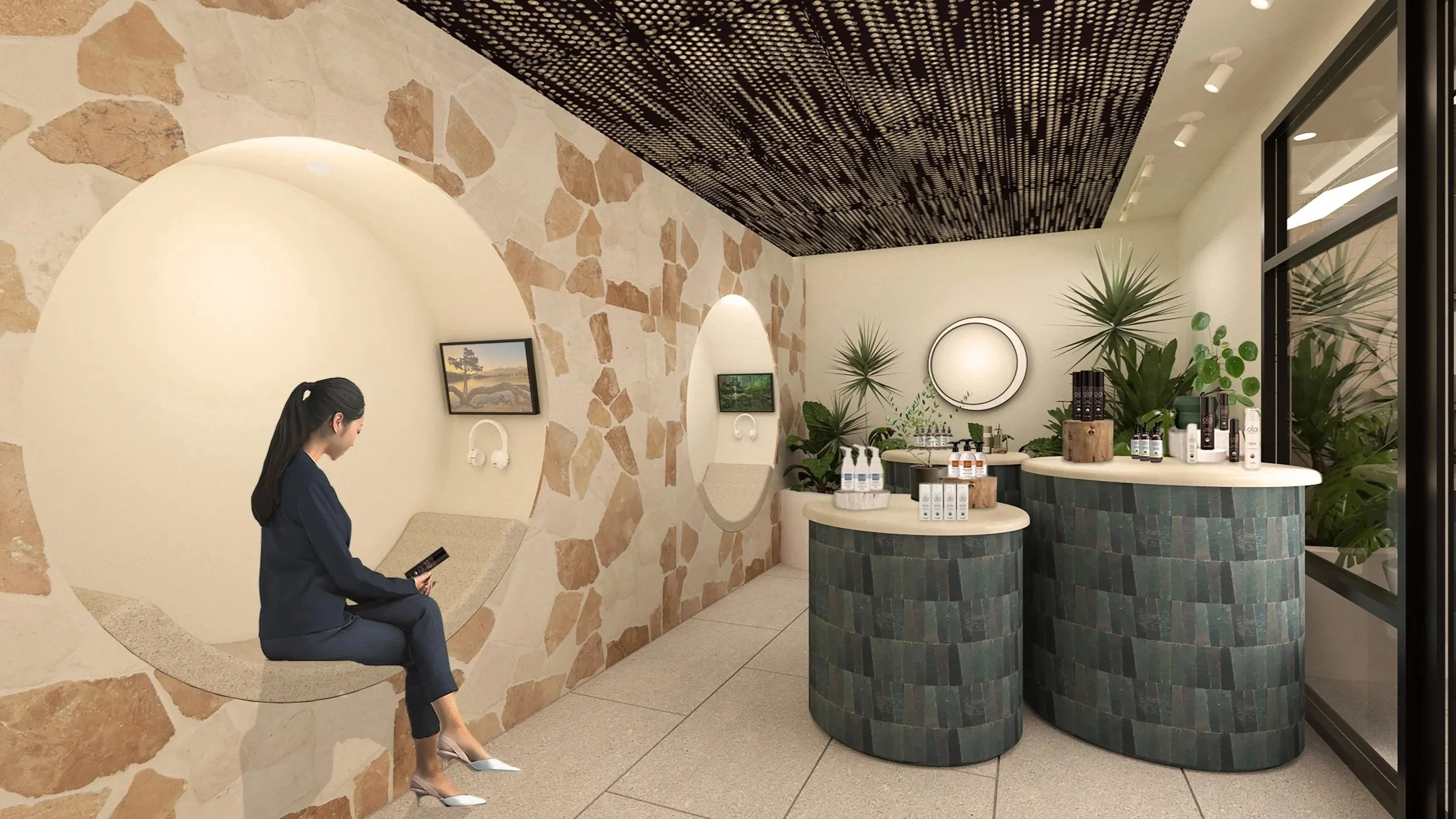 A woman with dark hair tied back, wearing a navy blue suit, sitting on a curved stone bench in a modern spa or wellness center. The room has a stone and white wall with artwork, built-in lighting, and lush green plants near a white circular mirror. There are two dark, tiled, semi-circular counters with bottles of skincare or wellness products on top, next to large windows.