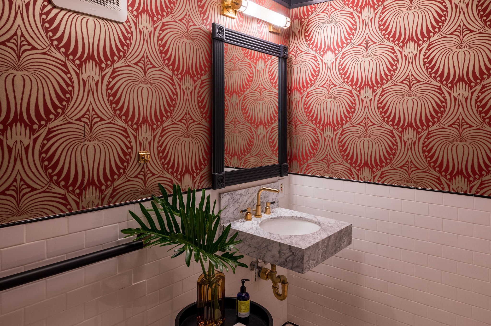 A bathroom wall decorated with red and gold floral-patterned wallpaper. Below the wallpaper, there is a white subway tile wall with black trim. A marble vanity with a white sink and gold faucet is in front of the wall. To the left, a green plant in a glass vase is visible, and perched on a black circular table, there are two bottles, one amber and the other blue. On the wall above the vanity, there is a large black framed mirror, and around it, a gold light fixture and a rectangular white light are mounted.