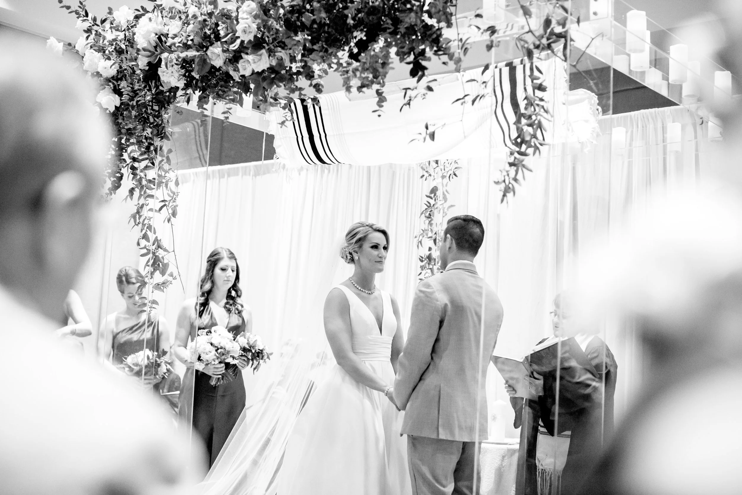 A black-and-white photo of a wedding ceremony where a couple is exchanging vows. The bride is in a white dress and the groom is in a suit. There are women standing behind the bride, and the setting is decorated with flowers and drapes.