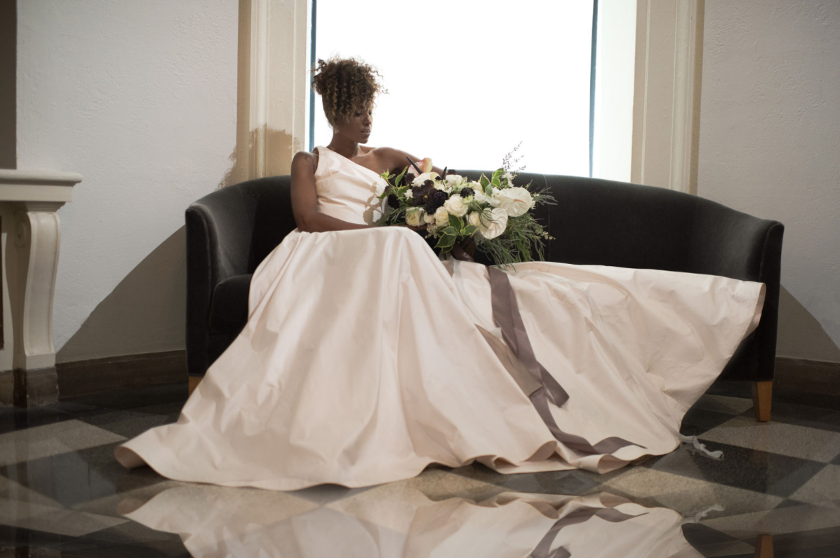A woman in a white dress sits on a black sofa holding a large bouquet of white and purple flowers, with a gray ribbon, in front of a window with bright light.