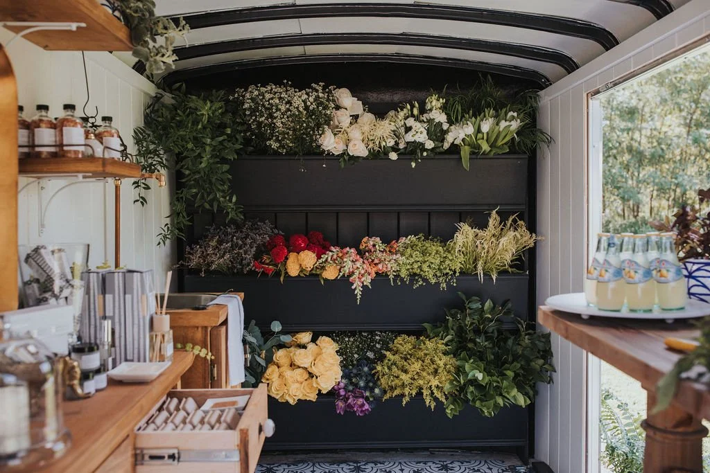 Indoor flower display with various flowers on black shelves and beverages on a counter, bright natural lighting.