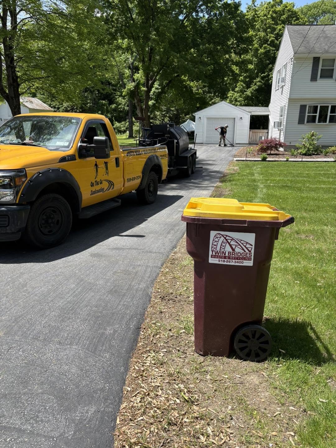A driveway with a yellow sanitation pickup truck parked on the left. A worker is near a garage in the background, and a maroon waste recycling bin with yellow lid is in the foreground. The house is white with gray shutters, surrounded by green trees and lawn.