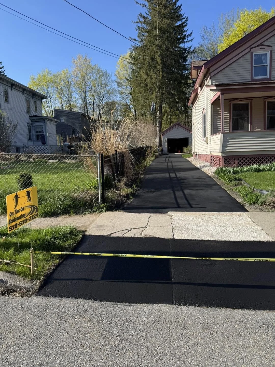 A freshly paved driveway leading to a garage, with a caution tape across the entrance and a yellow sign on the left indicating driveways are being resurfaced.
