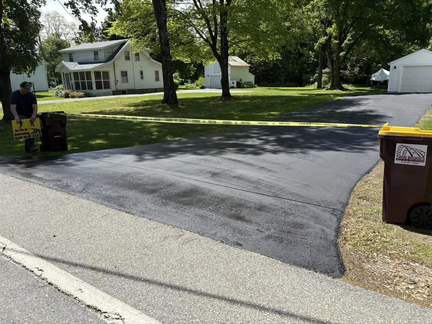 A newly paved driveway with yellow caution tape across it, a person installing or inspecting trim or bricks on the edge, a trash bin on the right, and residential houses and trees in the background.