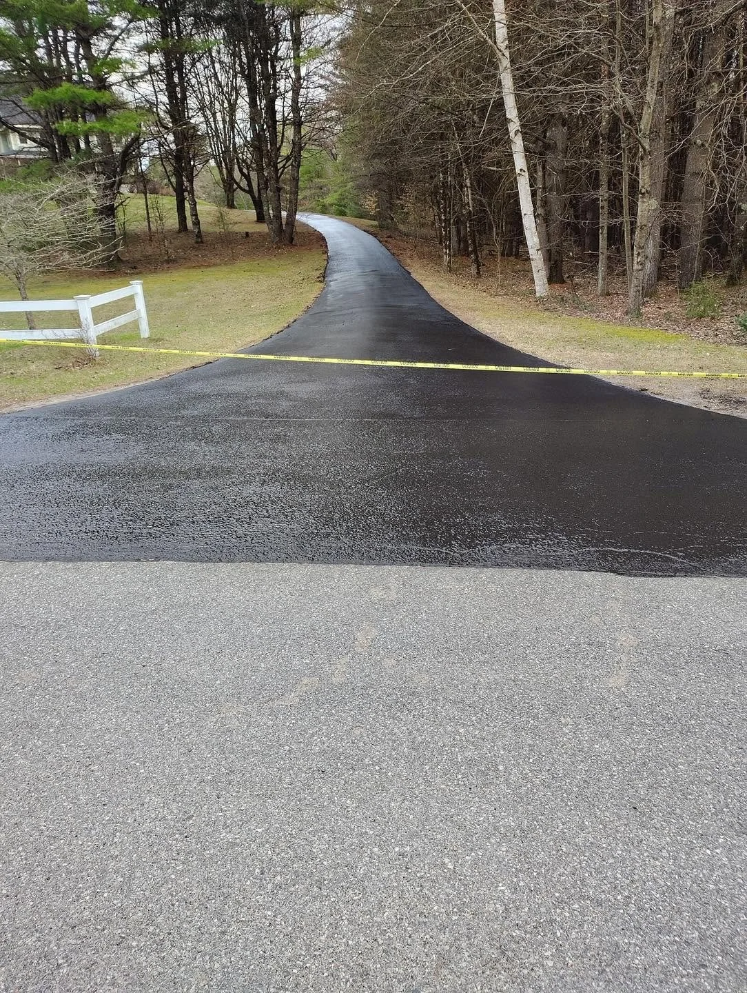 A newly paved black asphalt driveway curve leads into a wooded area, marked off with yellow caution tape.