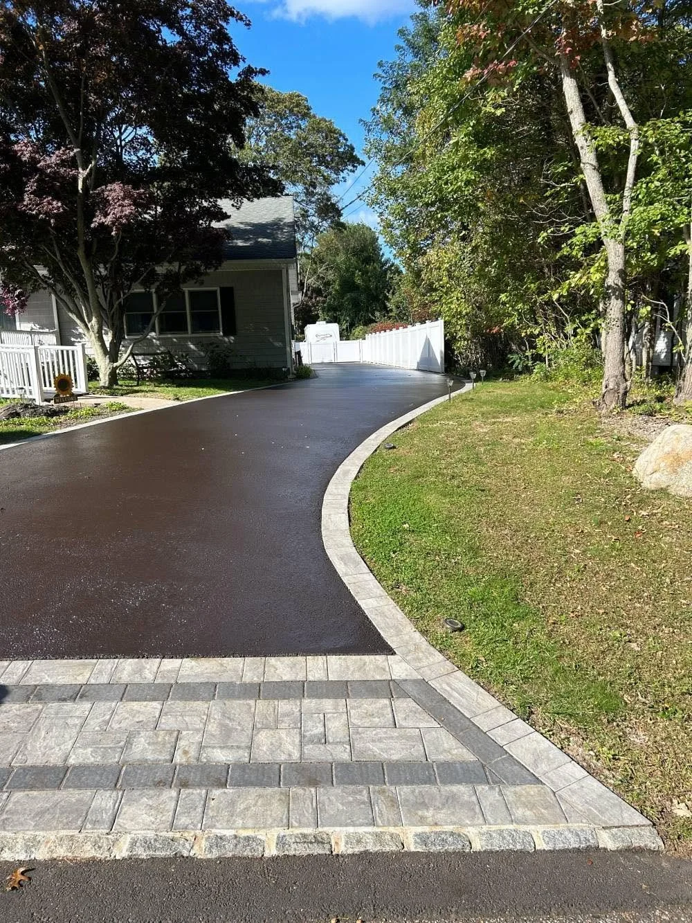 A freshly paved driveway curves past a house with a front porch, surrounded by trees and greenery, with a white fence and a blue sky overhead.