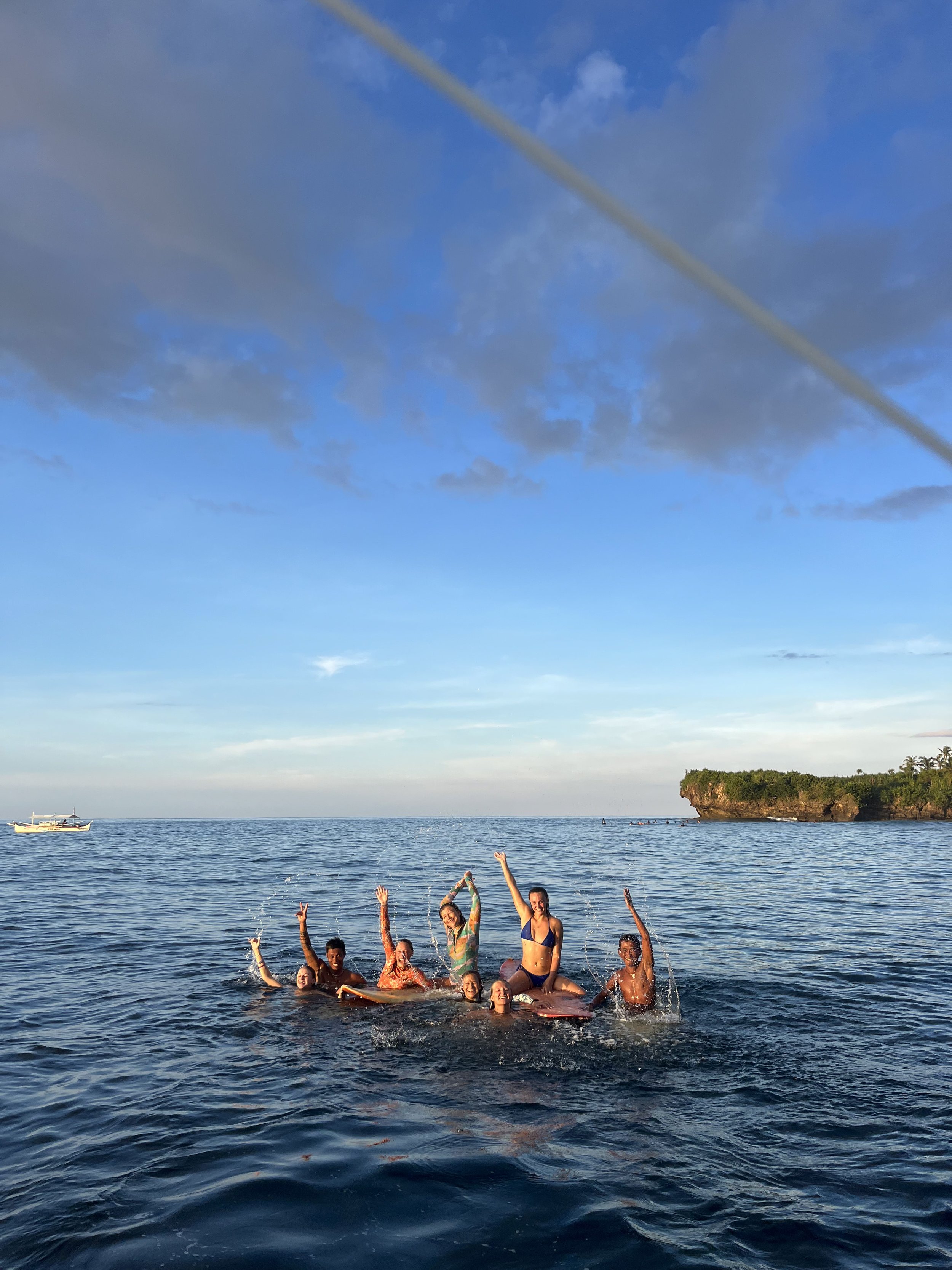 Group of people who Lara met during a trip, joyfully playing and splashing in the ocean near a rocky island in the philippines with greenery under a blue sky with clouds.