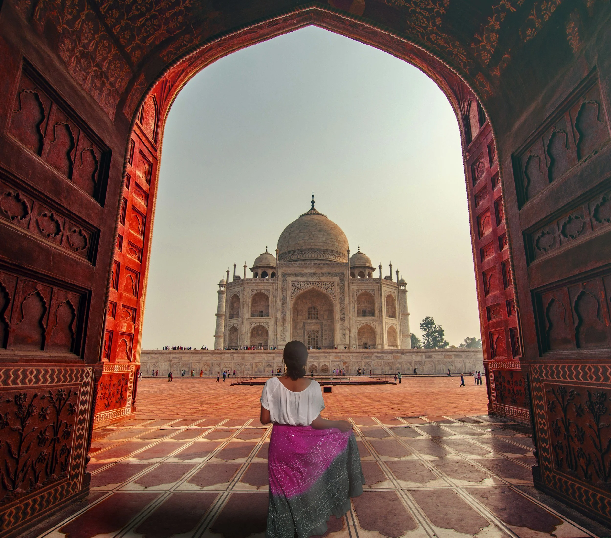 A young girl in a pink and gray traditional dress sitting on the floor inside a red sandstone archway with intricate carvings, looking at the Taj Mahal in India through the arch.