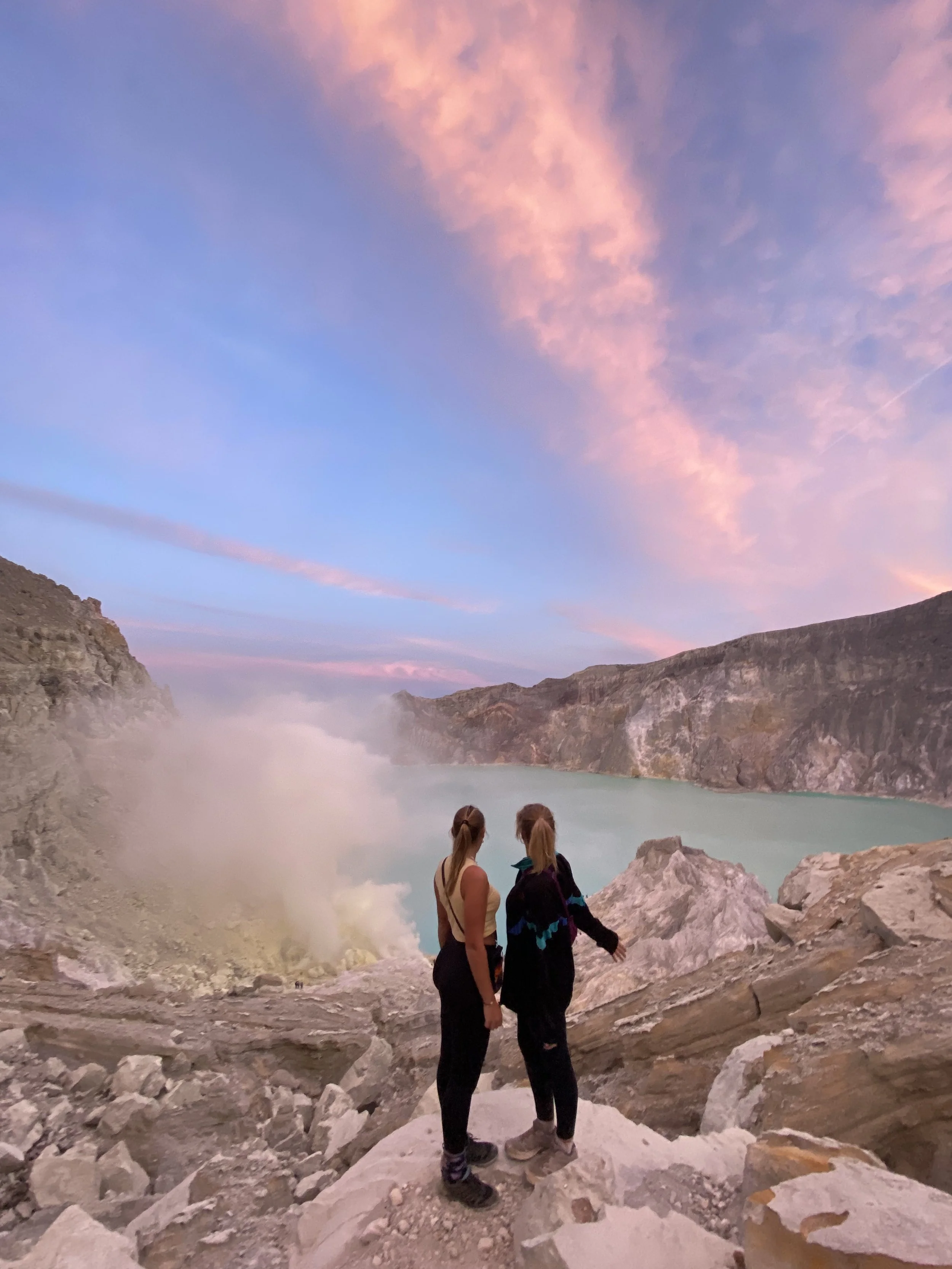 Lara and Danique standing on rocky terrain near a large body of water, with cliffs on either side, under a colorful sky with pink and purple clouds at Mount Ijen in Indonesia.