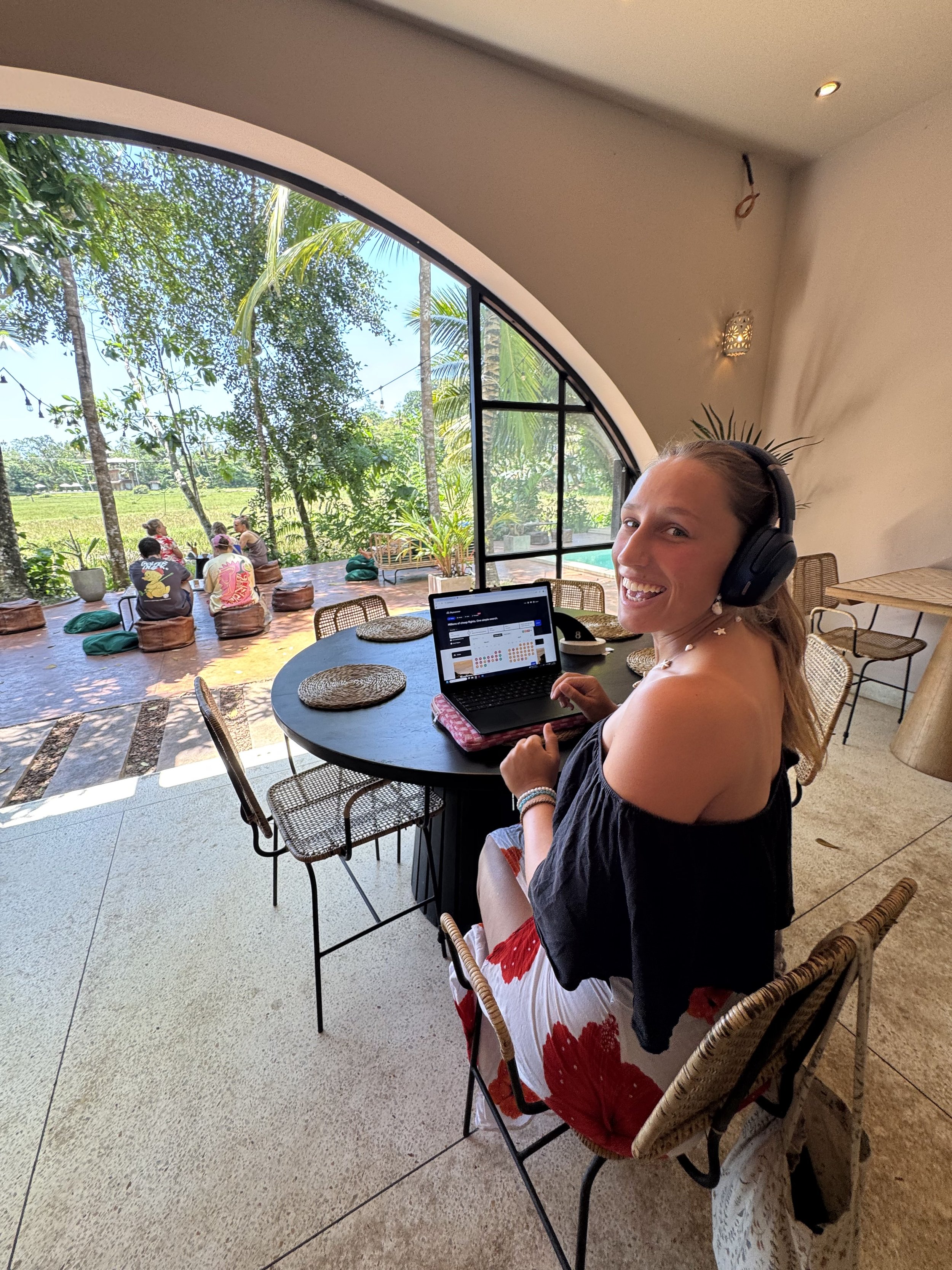 Lara smiling wearing headphones sitting at a table with a laptop in a cafe with large window showing outdoor green trees and people seated outside in Ahangama, Sri Lanka.