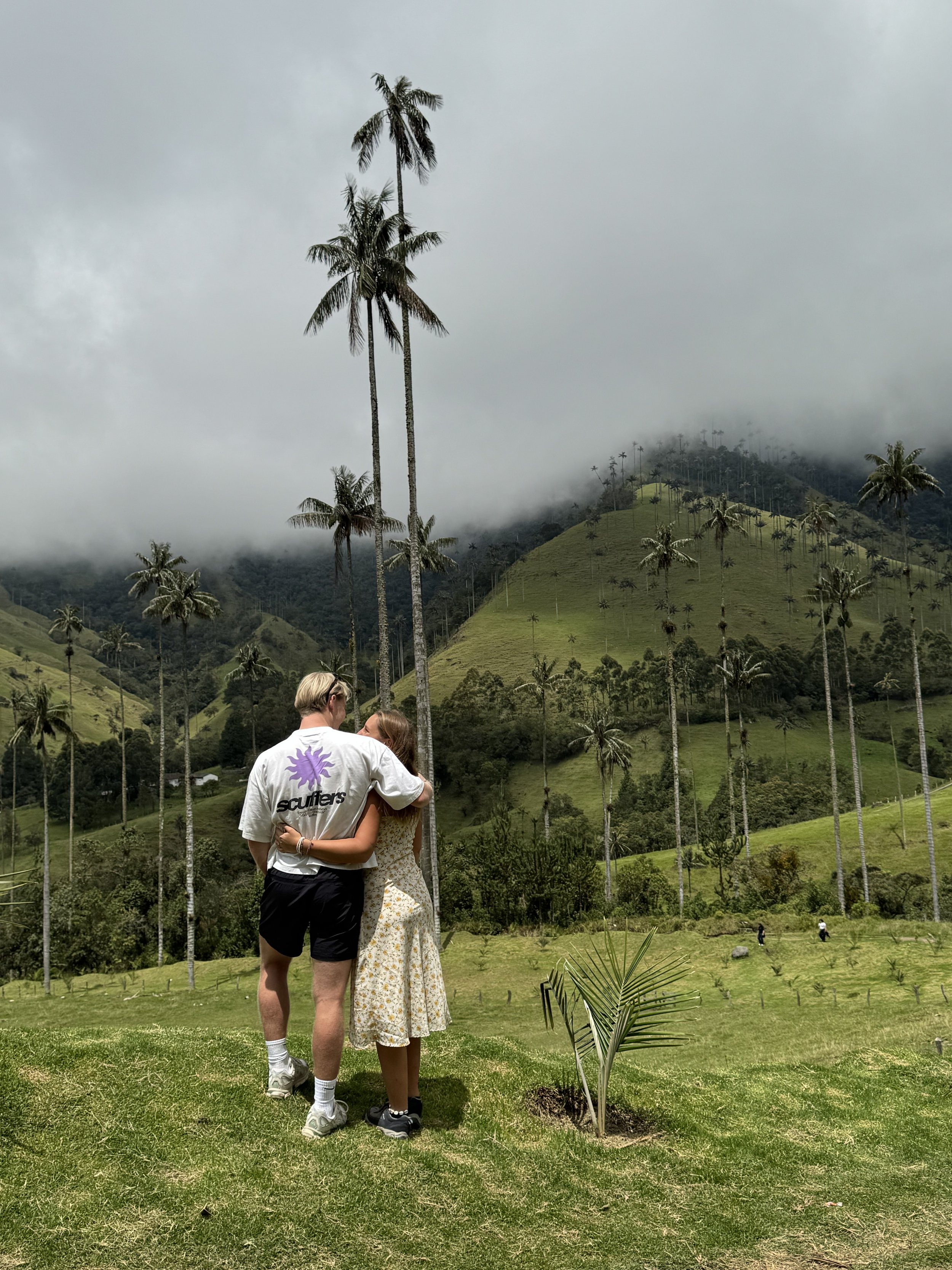 Lara and Jonas embraced and looking at each other in The Coccora Valley near Salento in Colombia with tall palm trees, rolling hills, fog, and mountains in the background.