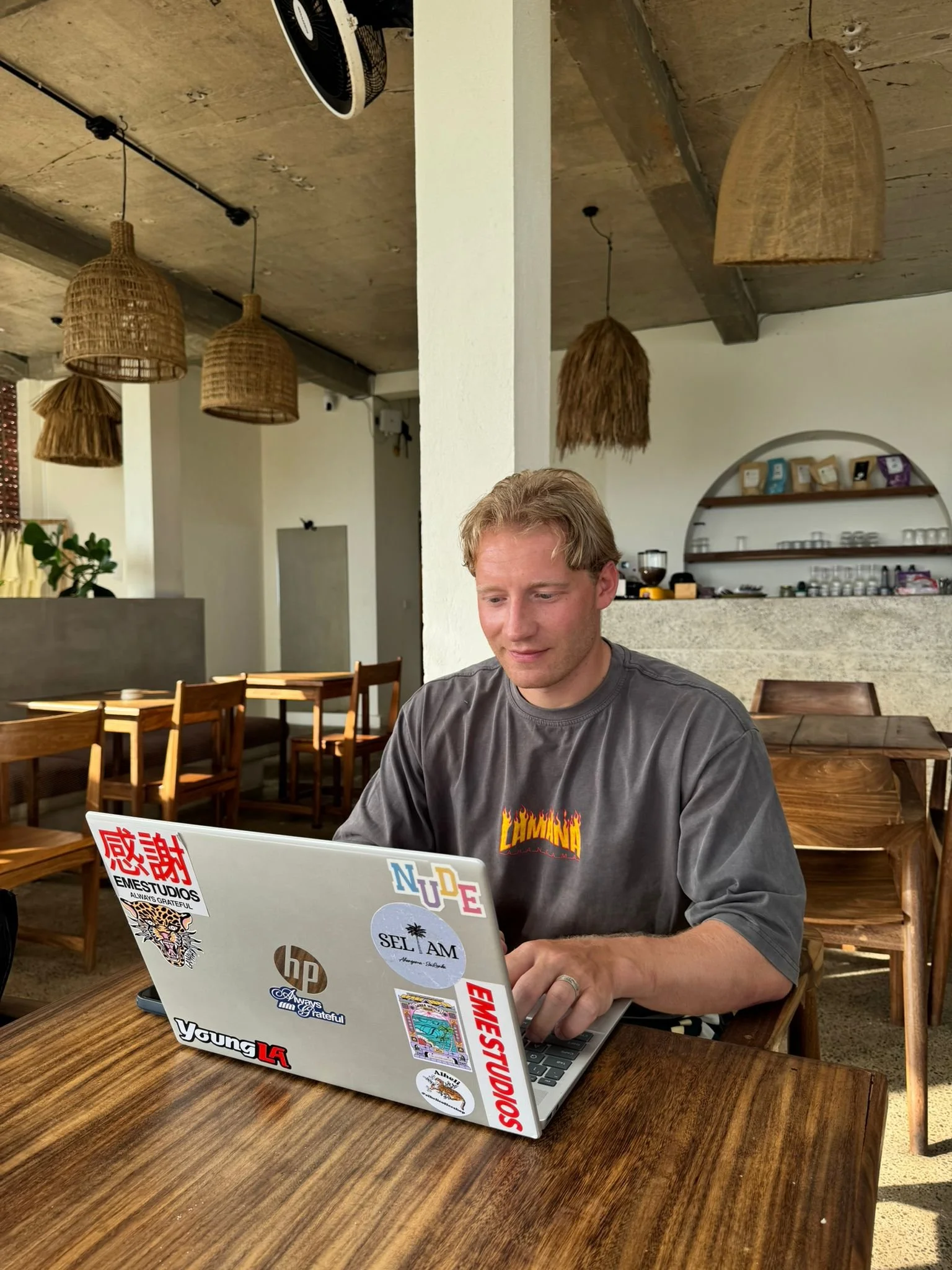 Jonas working on his laptop at a wooden table in a cafe, with a white wall, wooden chairs, modern hanging wicker lamps, and shelves with coffee bags and equipment in the background in Ahangama, Sri Lanka. the café is named Teddies.