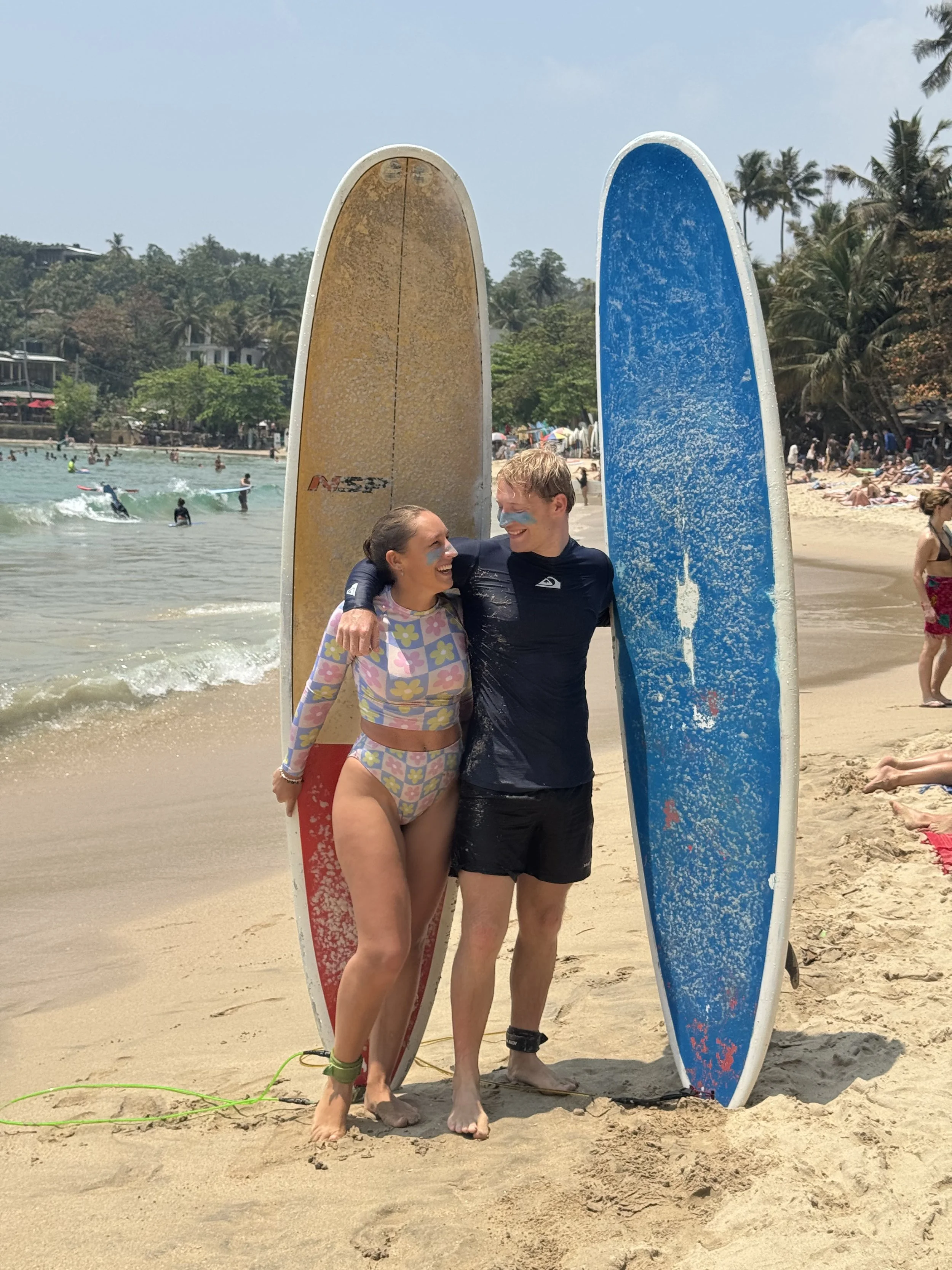 Lara and Jonas stand on a sandy beach holding surfboards, smiling and looking at each other. In the background, there are surfers in the water and people laying on the beach in Hiriketiya, Sri Lanka