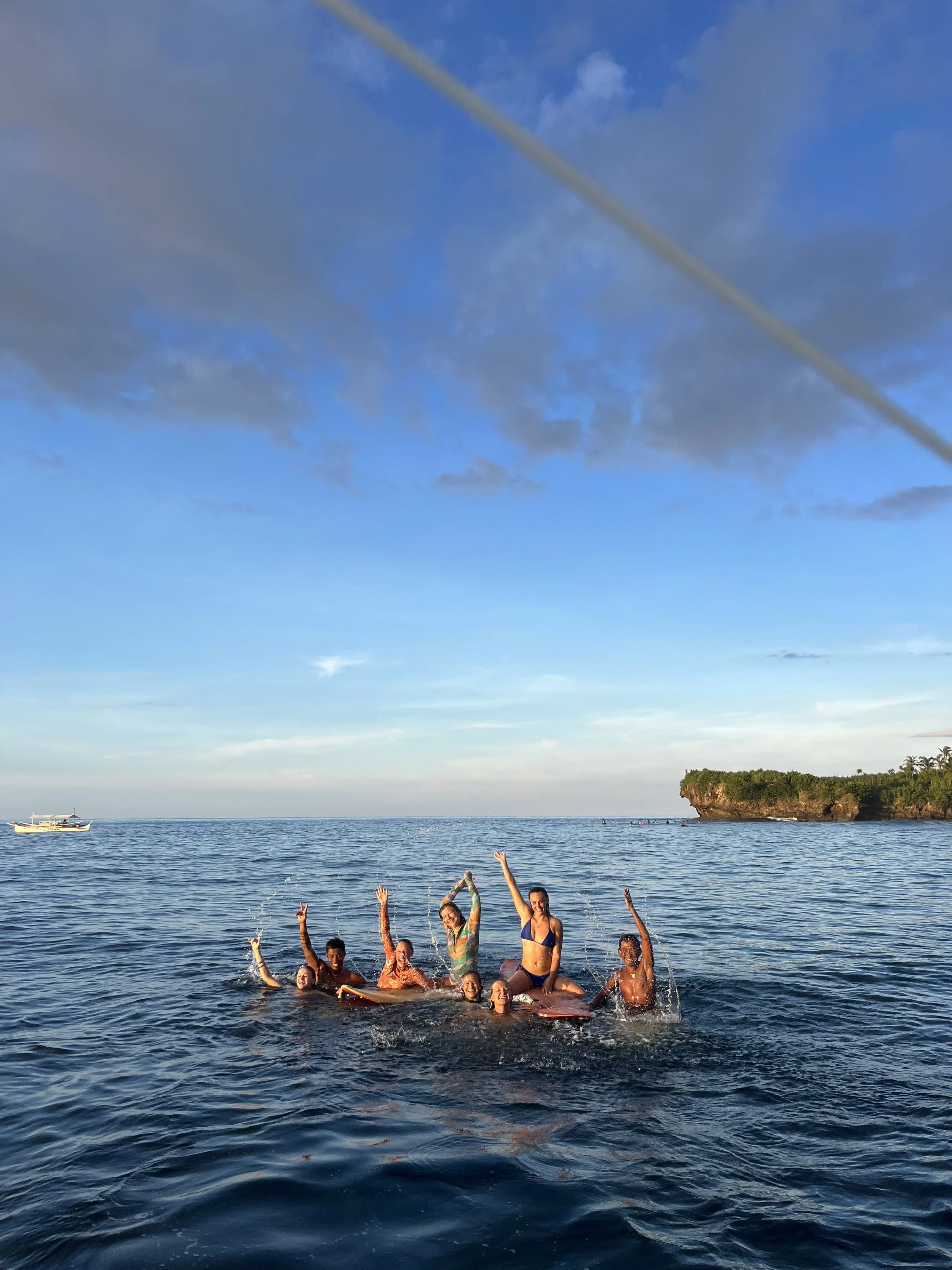 Group of people who Lara met during a trip, joyfully playing and splashing in the ocean near a rocky island in the philippines with greenery under a blue sky with clouds.