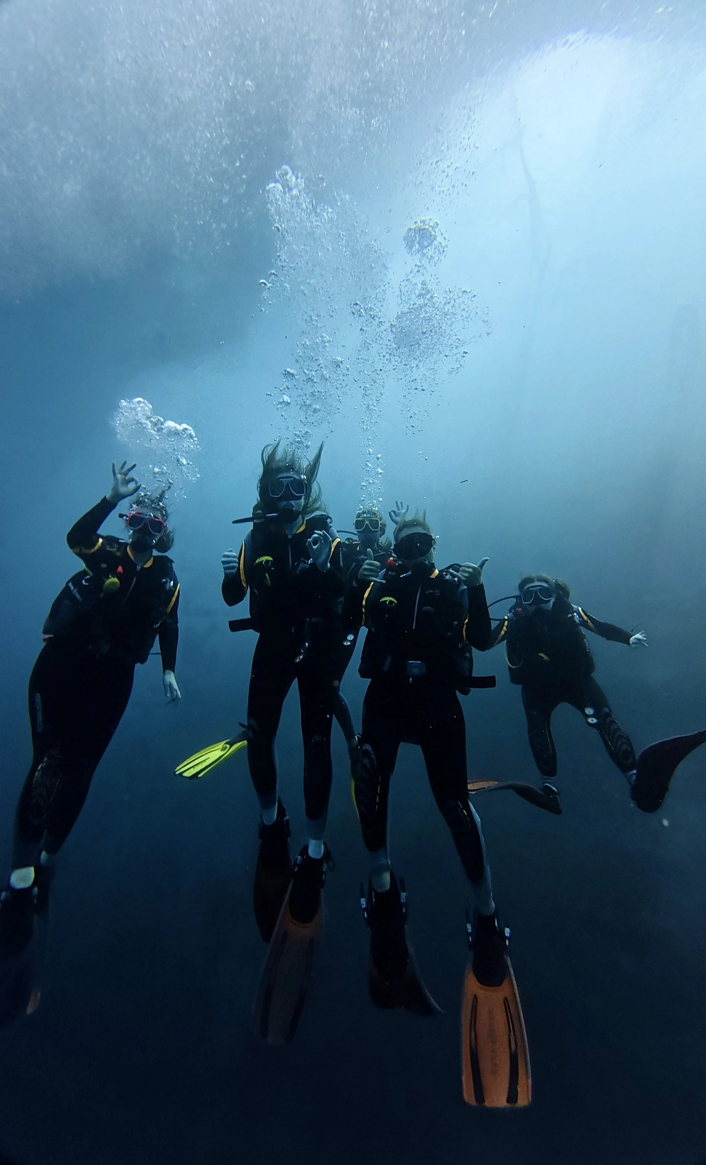 Four scuba divers underwater, wearing wetsuits, masks, and fins, making hand gestures, with bubbles rising above. the lighting indicates they are in open water in the maldives.