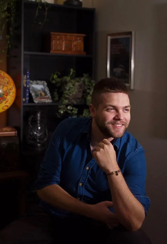 A young man with short brown hair and a beard, wearing a blue button-down shirt, sitting indoors with a thoughtful smile, resting his chin on his hand, in a cozy room with shelves, plants, and a globe in the background.