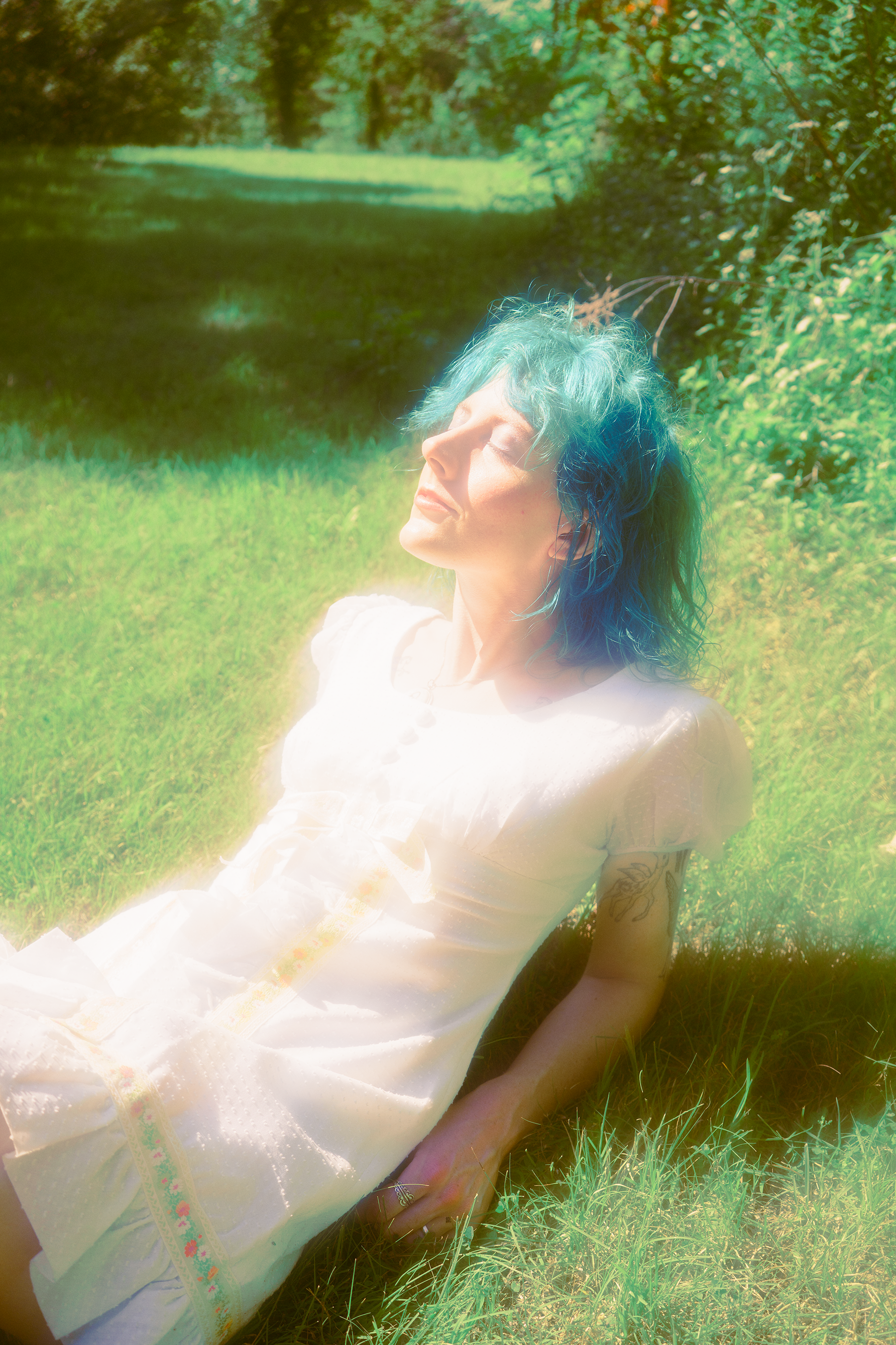 Artist Jonie laying on the grass in a white sundress, eyes are closed, with face turned towards the sun, resting on her elbows, her hair is a blue green color.