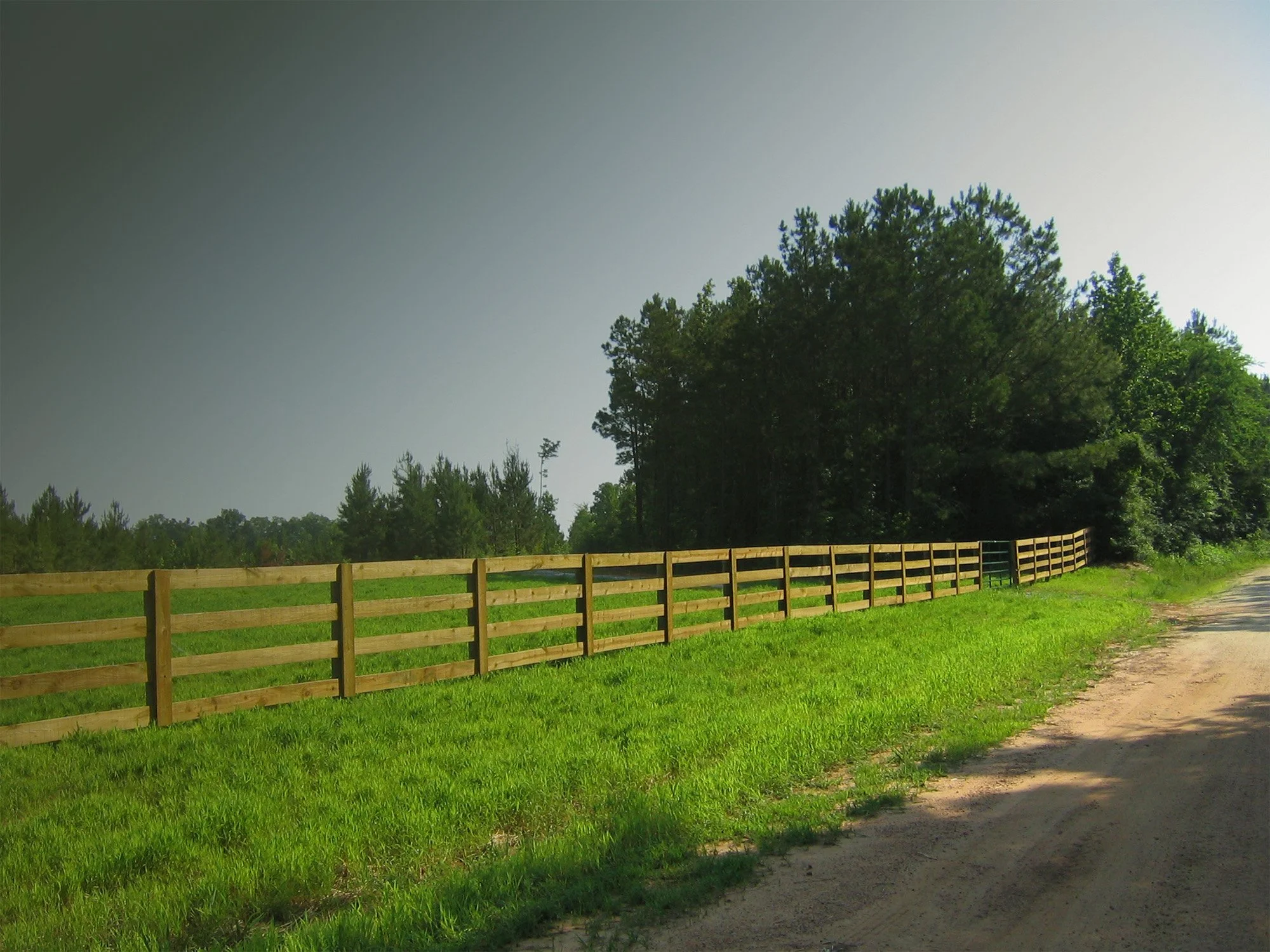 A rural landscape with a dirt path on the right, a wooden fence parallel to it, green grass, trees in the background, and a cloudy sky.