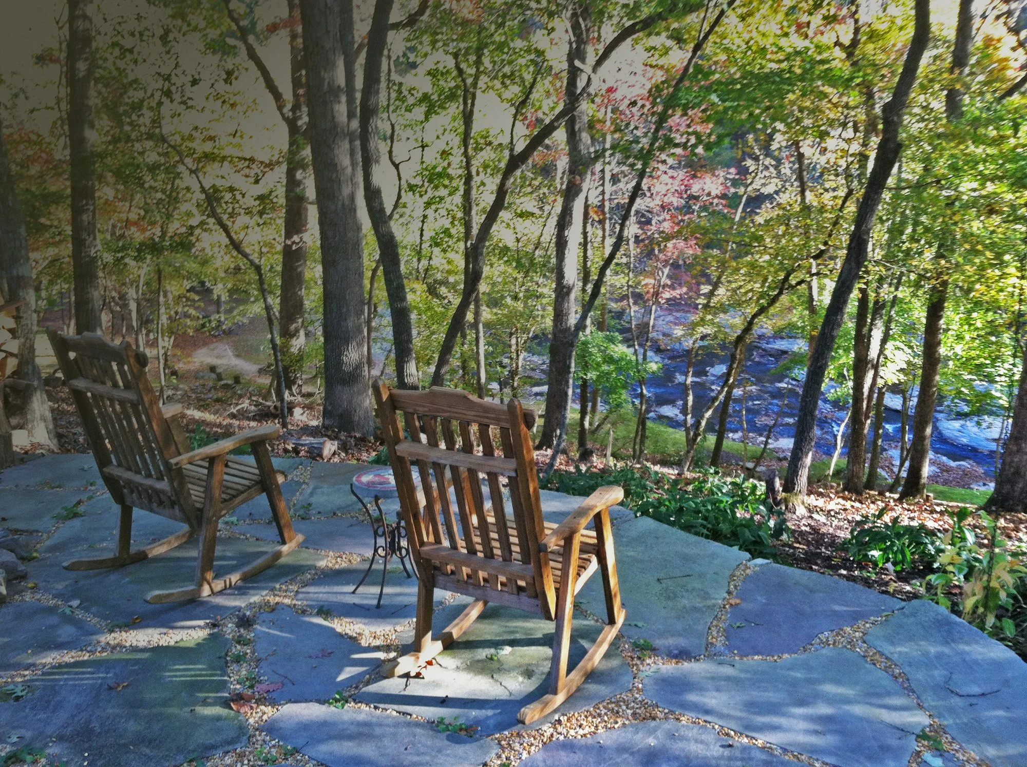 A backyard patio with wooden chairs, surrounded by trees with green and autumn-colored leaves, and a forested area with a creek in the background.