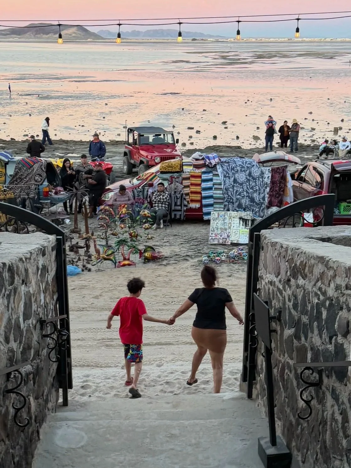 View of a beach scene at sunset with vendors selling colorful blankets, clothing, and crafts near the shore, and people walking along the beach, with mountains in the background. Two people, a woman and a child, are holding hands at the entrance, walking towards the beach.