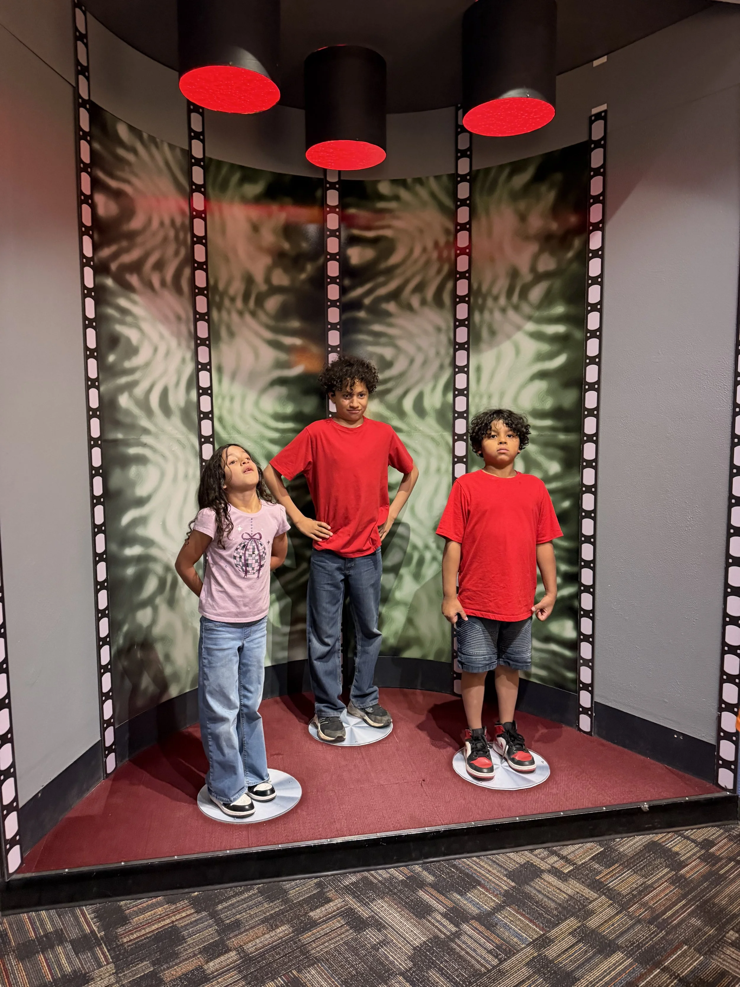 Three children standing on a platform with a film strip themed backdrop and spotlight on each, inside a room with patterned carpet.