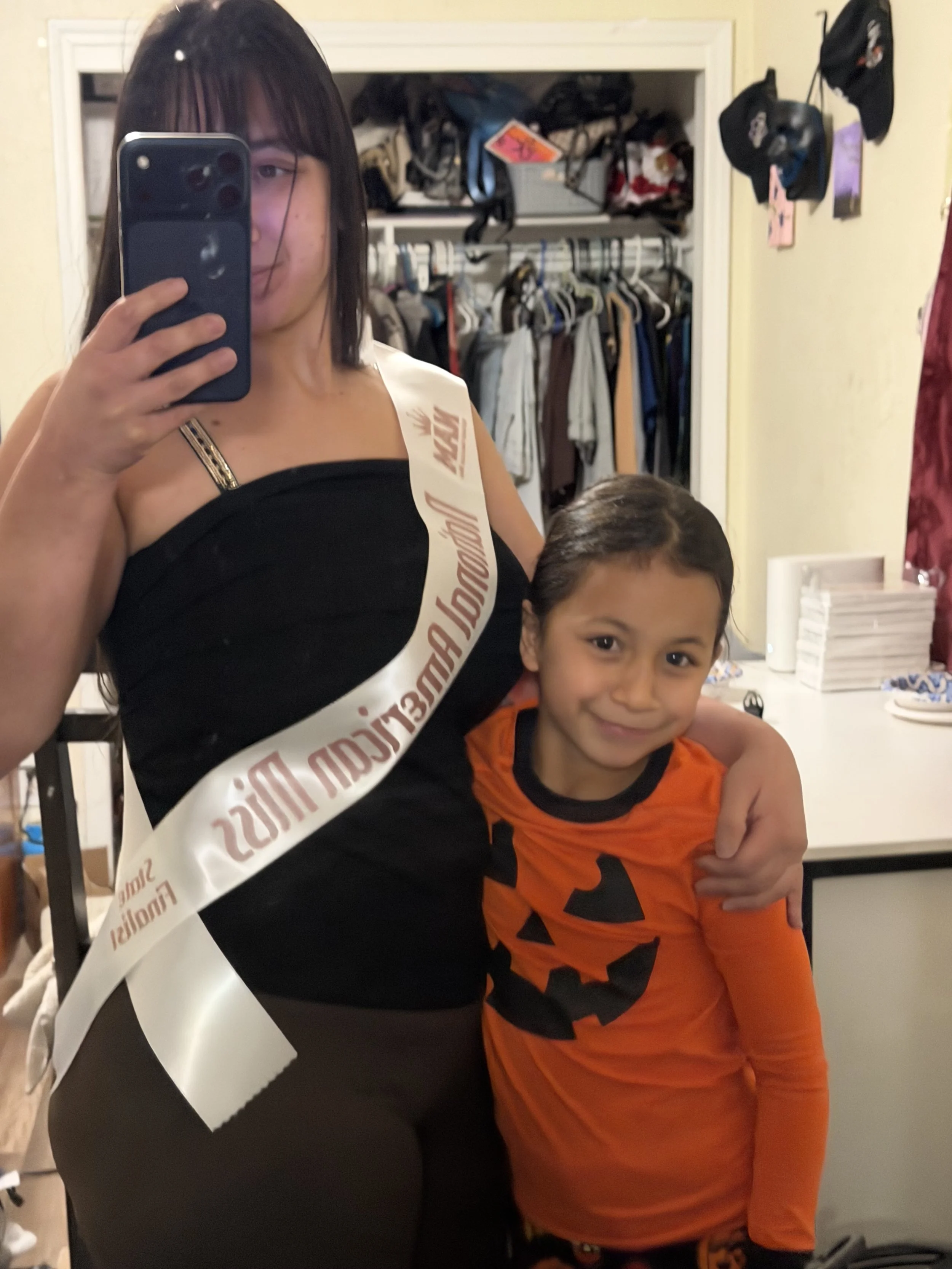 A woman taking a mirror selfie with a young girl in Halloween costume. The woman is wearing a sash and black clothing, and the little girl is dressed as a pumpkin.
