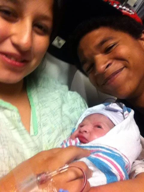 A woman and man smiling while holding a newborn baby wrapped in a hospital blanket.