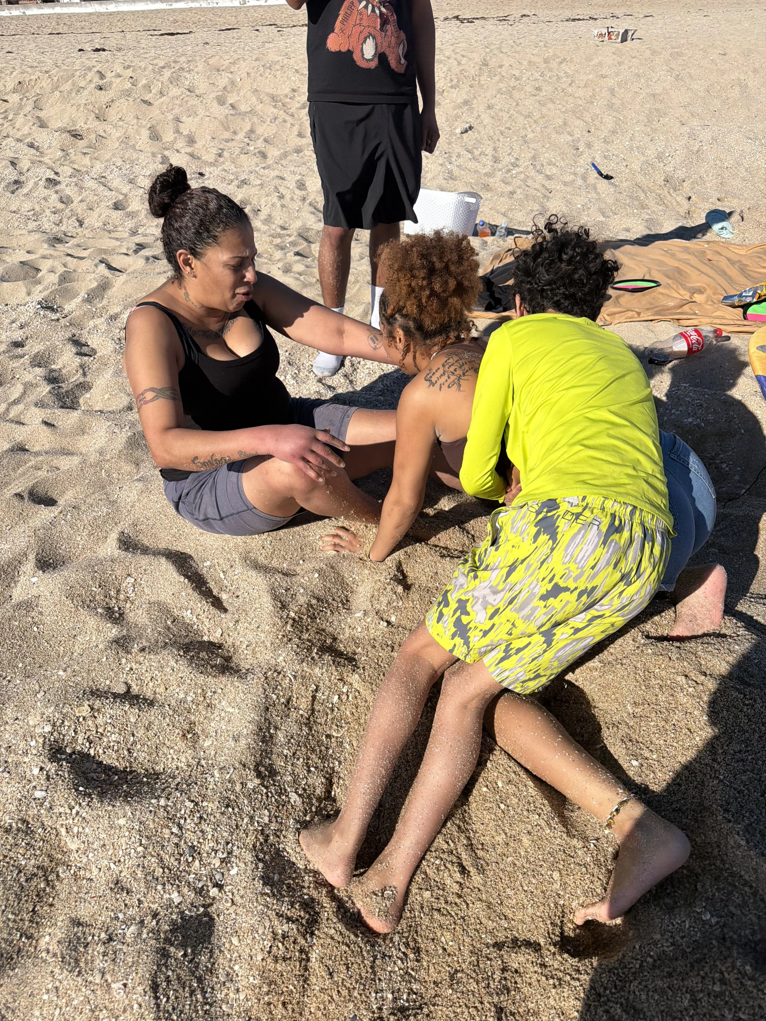 Three people are on a sandy beach, with two women kneeling on the sand and a man standing behind them. The women are holding hands and appear to be engaged in a moment of connection or prayer. The scene is sunny, and various beach items, including water bottles and a towel, are scattered around.
