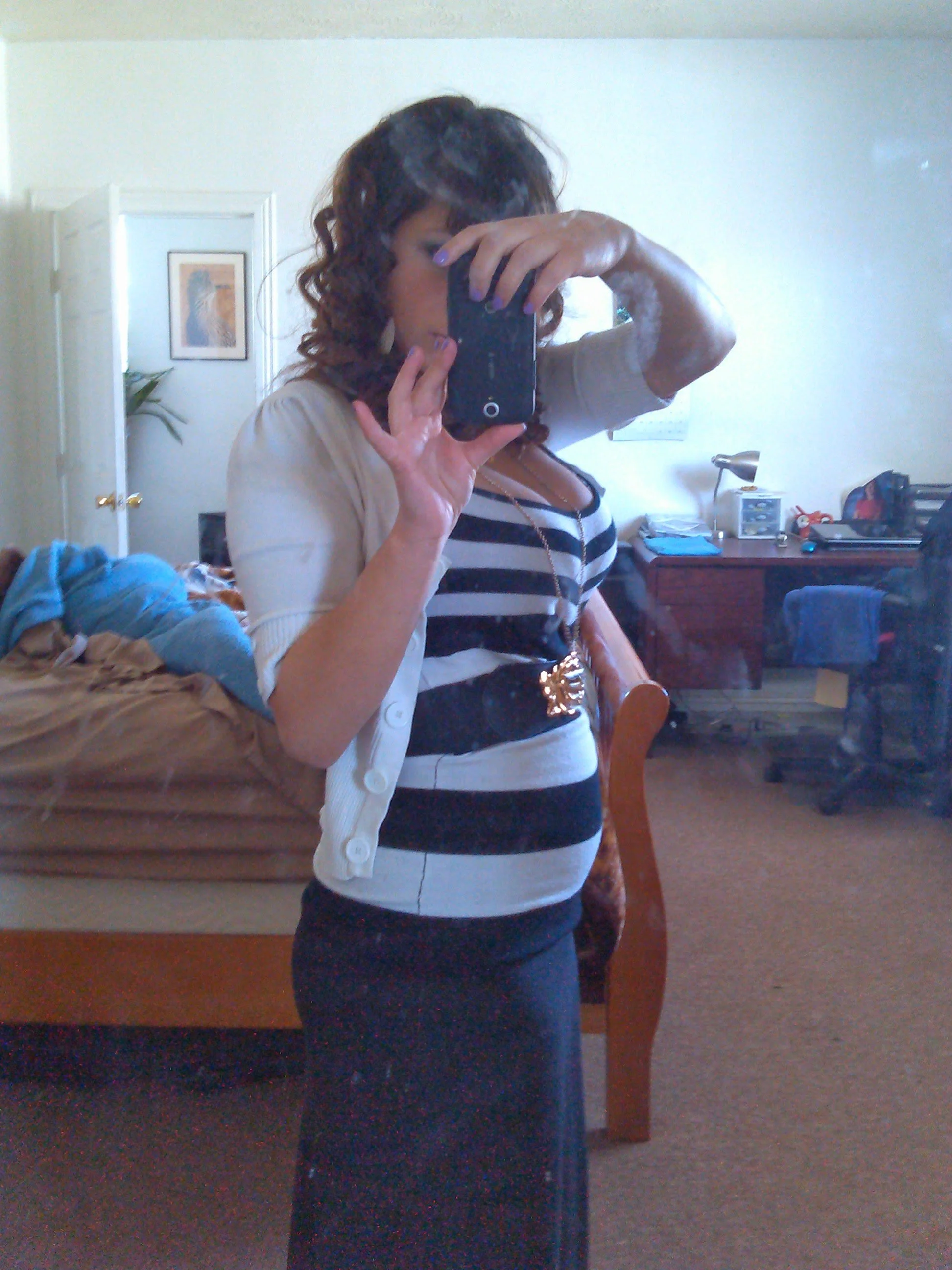 A woman with curly hair taking a mirror selfie in a bedroom. She is wearing a black and white striped top, a beige cardigan, and a gold belt. The room has a bed with laundry, a wooden dresser, and wall decorations.
