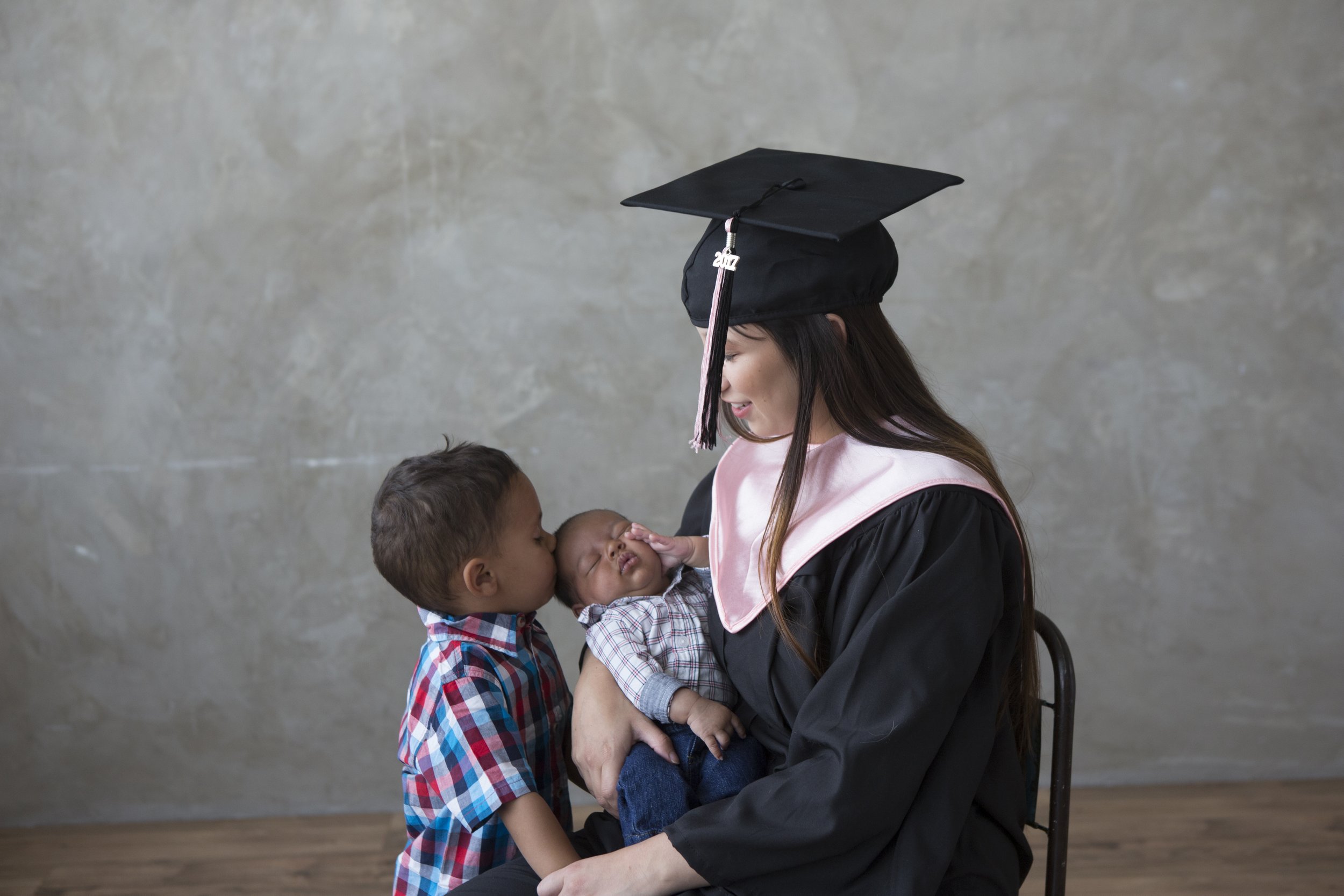 A woman in a graduation gown and cap holding a sleeping baby and two young children, sharing a kiss on the baby's cheek against a plain textured wall.