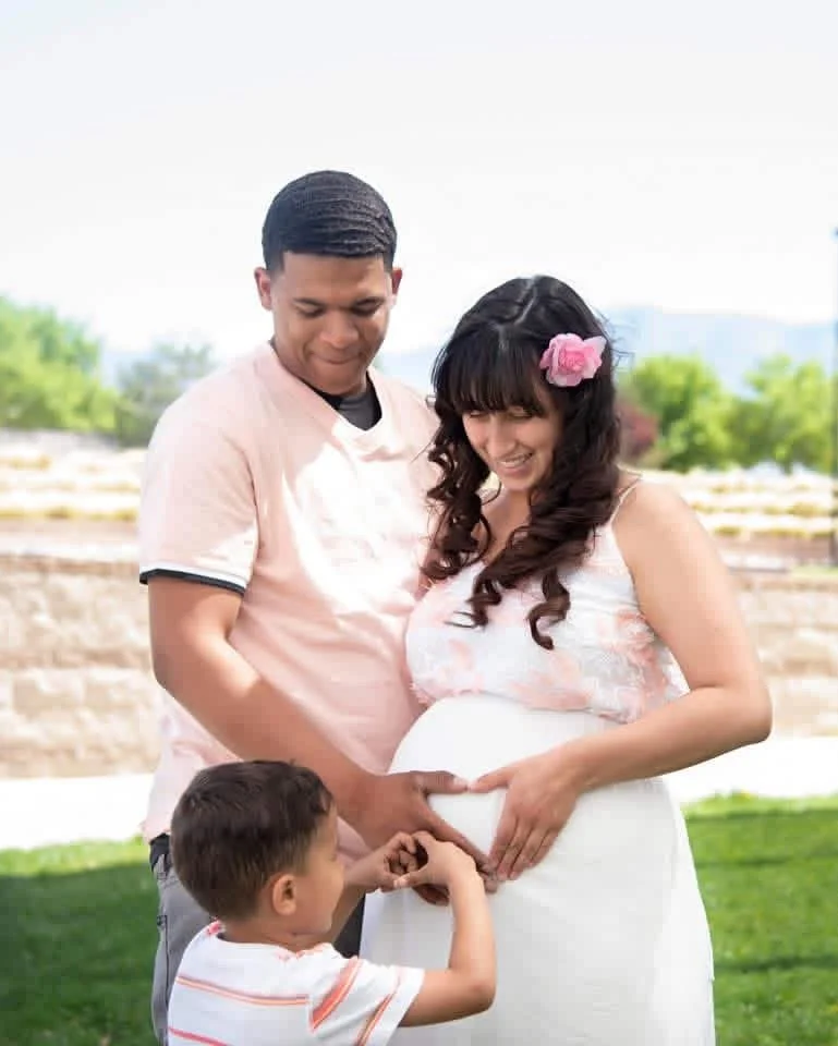A happy pregnant woman with dark, curly hair, wearing a pink flower hair accessory and a white dress, stands outdoors with her family. A man, possibly her partner, and a young boy, likely their son, touch her pregnant belly, which they are all smiling at. The background features greenery and a bright sky.