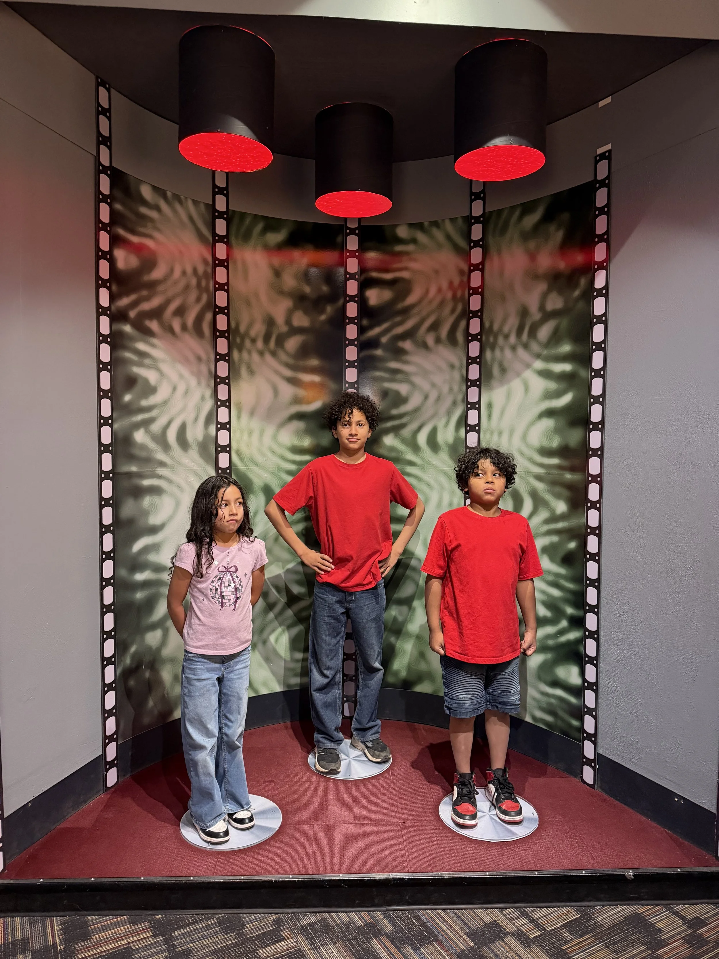 Three children standing on circular platforms in front of a green and black patterned backdrop, with red overhead lights and black and white film strip borders.